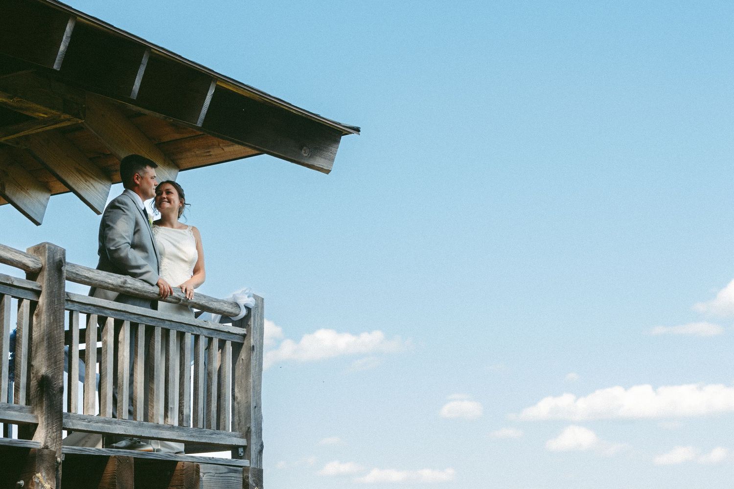 Couple standing on wooden deck overlooking scenic mountain landscape on sunny day.