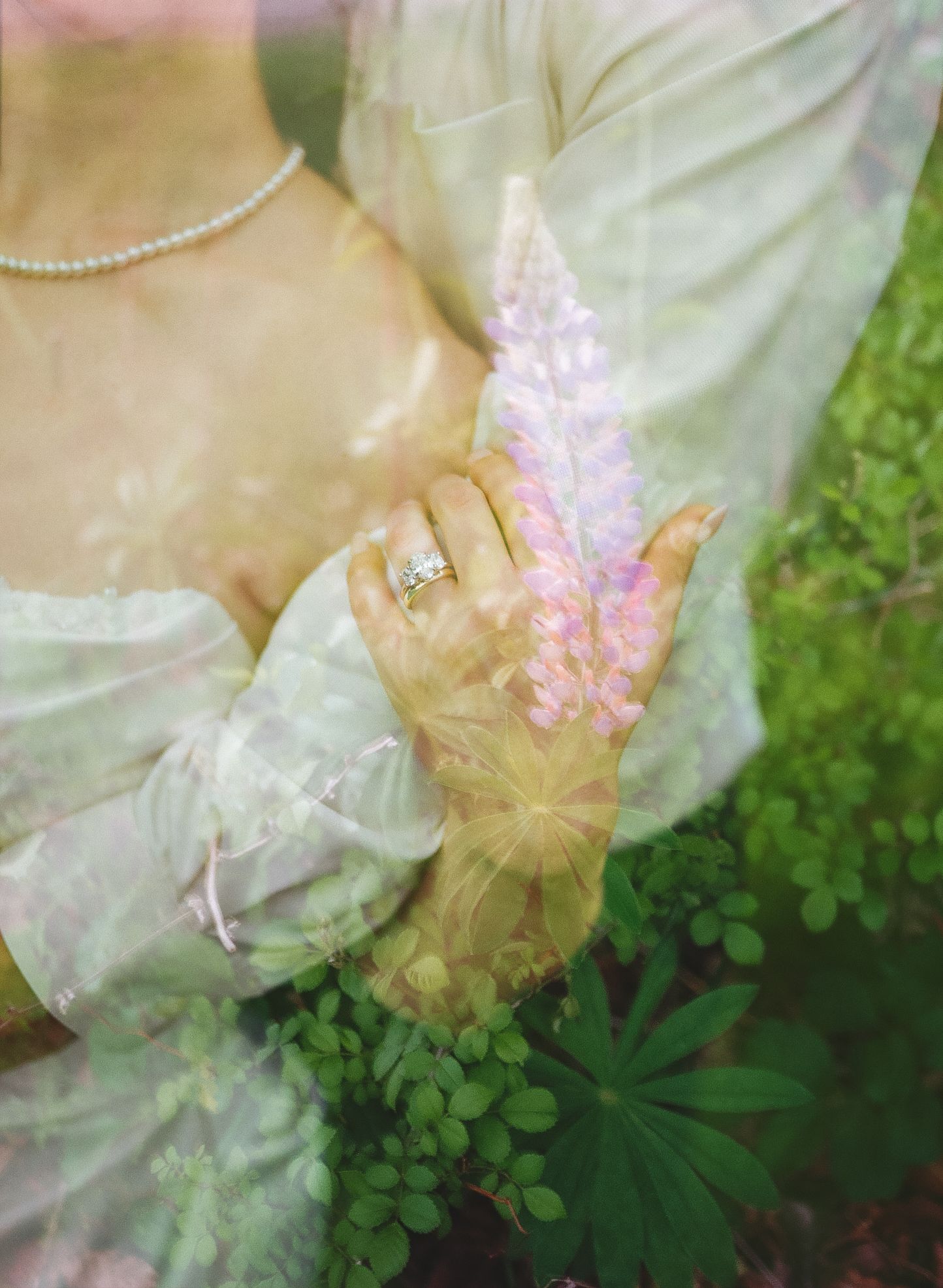 A bride in a sheer green dress holds a pink lupine flower, showcasing a diamond engagement ring and pearl necklace.