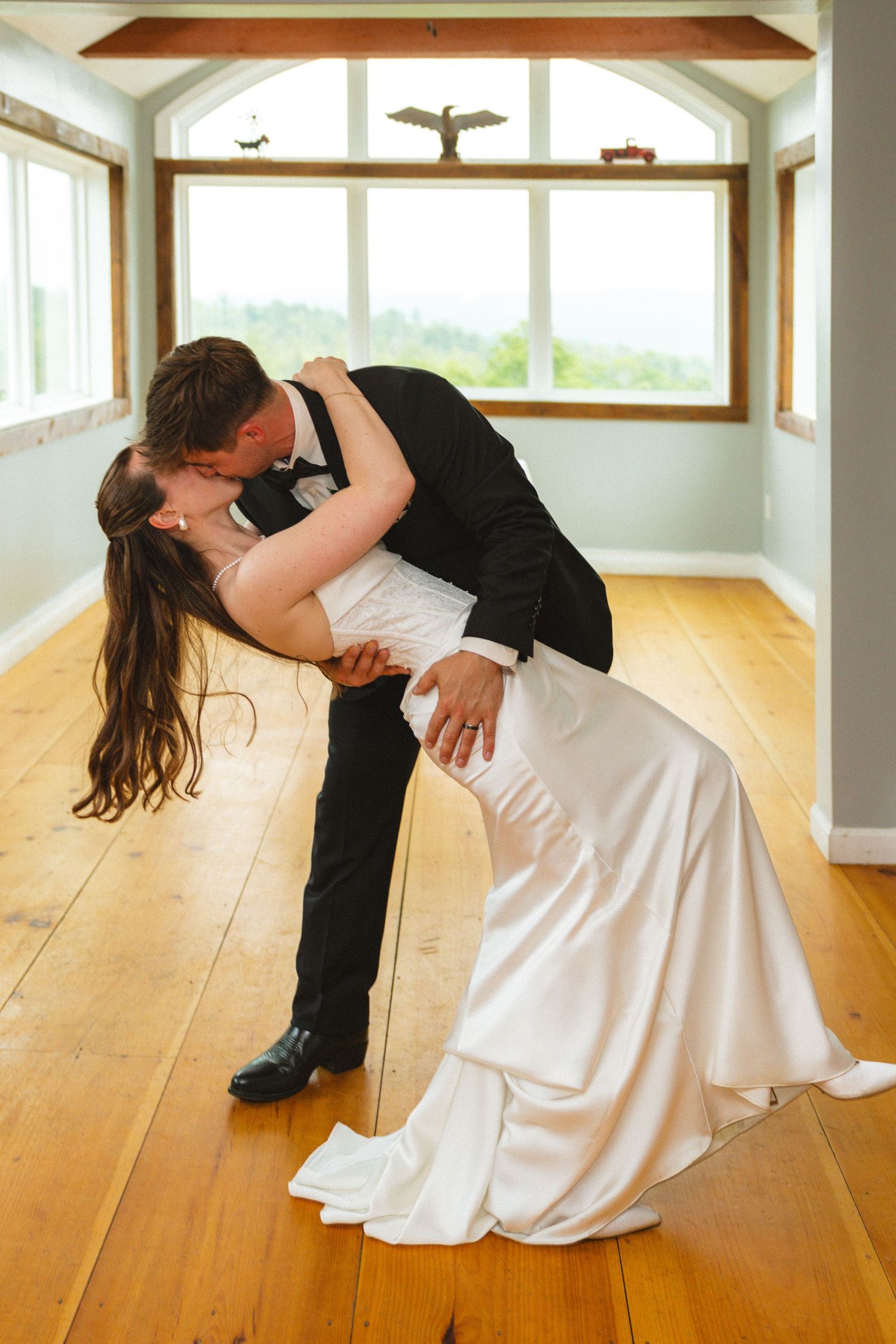 Bride and groom share a romantic dip kiss on hardwood floors in a bright, windowed room during their wedding day.