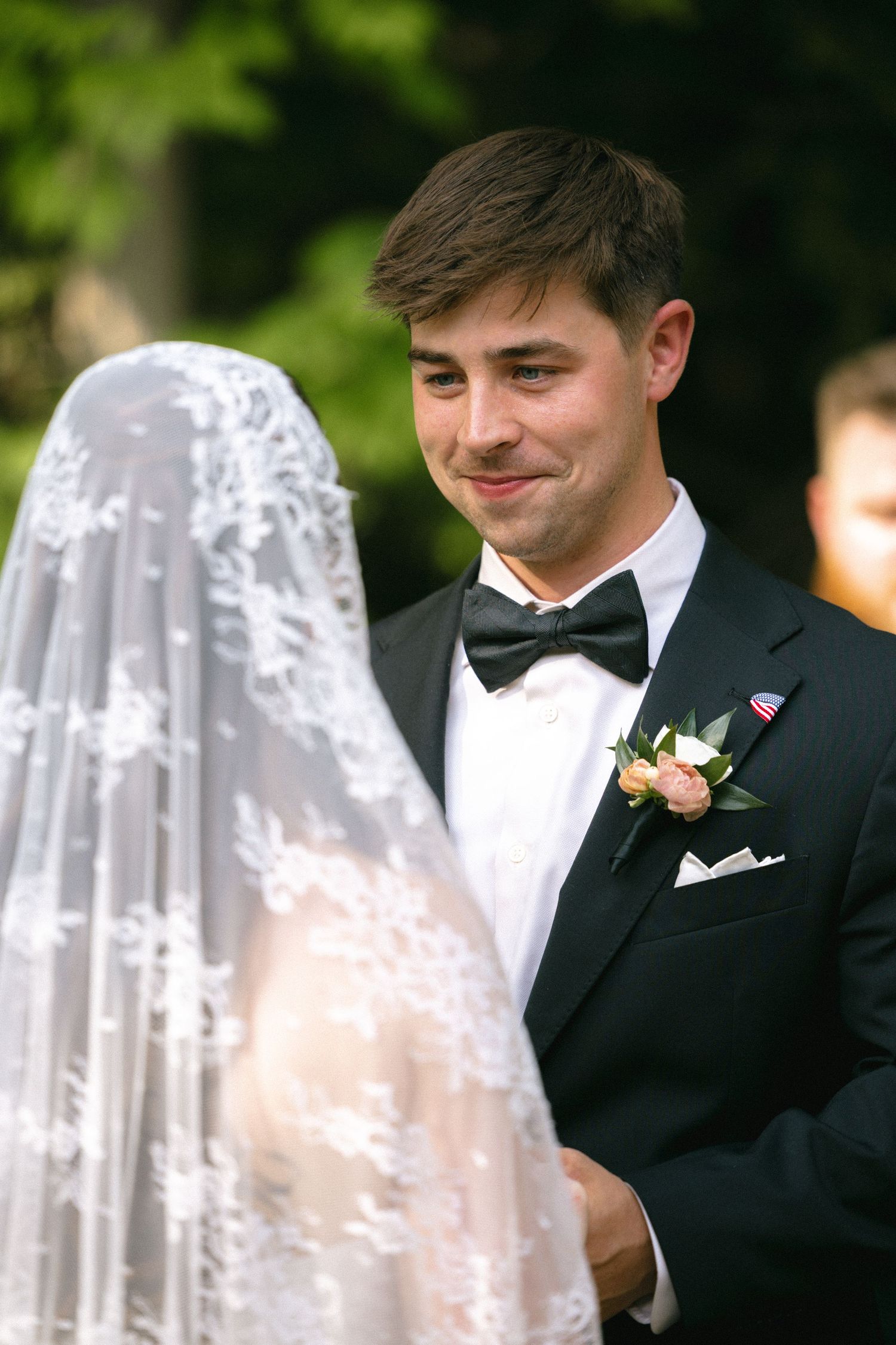 Groom in black tuxedo smiling tenderly at bride in lace veil during outdoor wedding ceremony.
