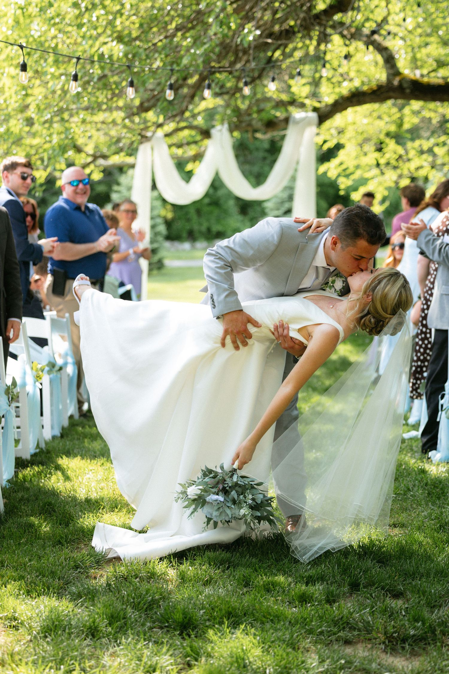 Groom dips bride for a kiss at outdoor wedding ceremony on green lawn with white chairs and draped arch.
