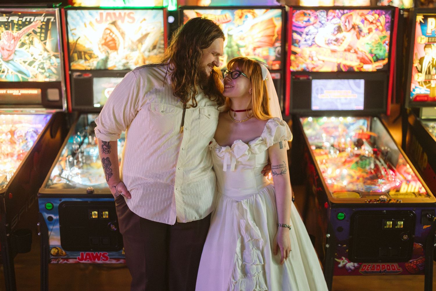 Couple in wedding attire sharing a romantic moment in front of colorful pinball machines at an arcade venue.