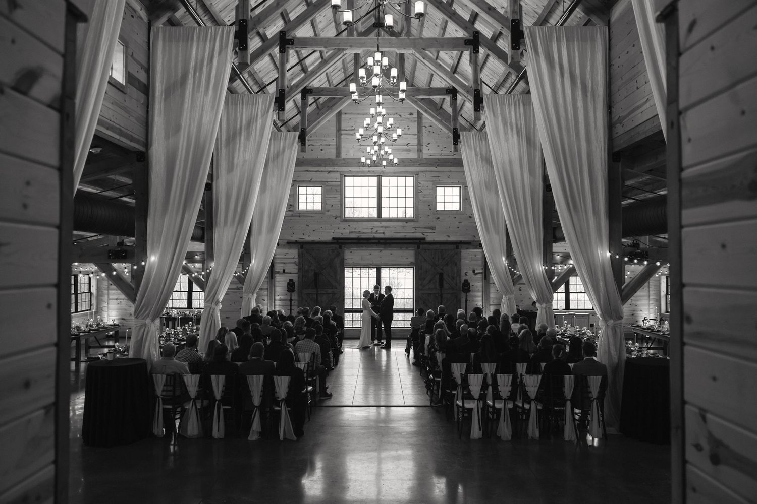 Black and white photograph of a wedding ceremony in a rustic barn venue with dramatic lighting and flowing white drapes.