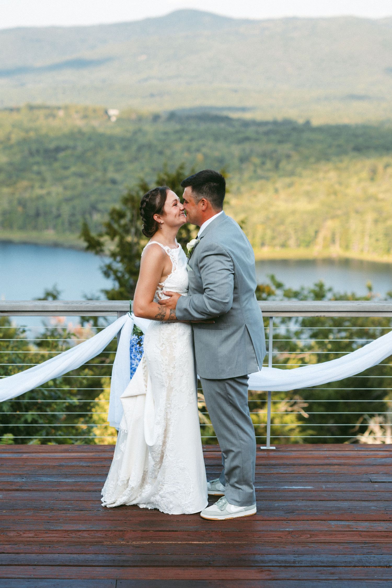 A couple shares a romantic moment on a deck overlooking mountains and a lake.