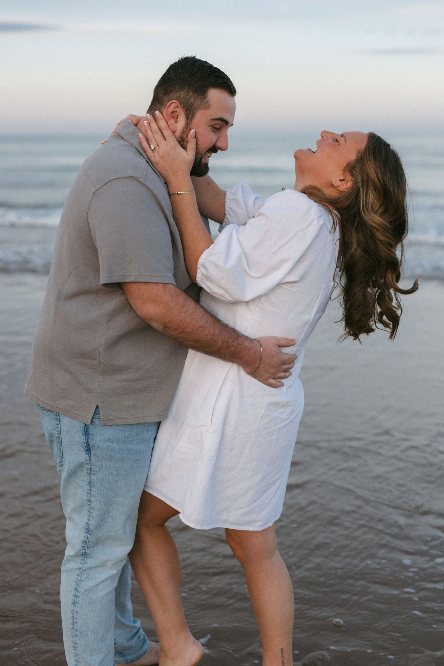 Couple embracing and laughing together on a beach at sunset, woman in white dress, man in gray shirt and jeans.