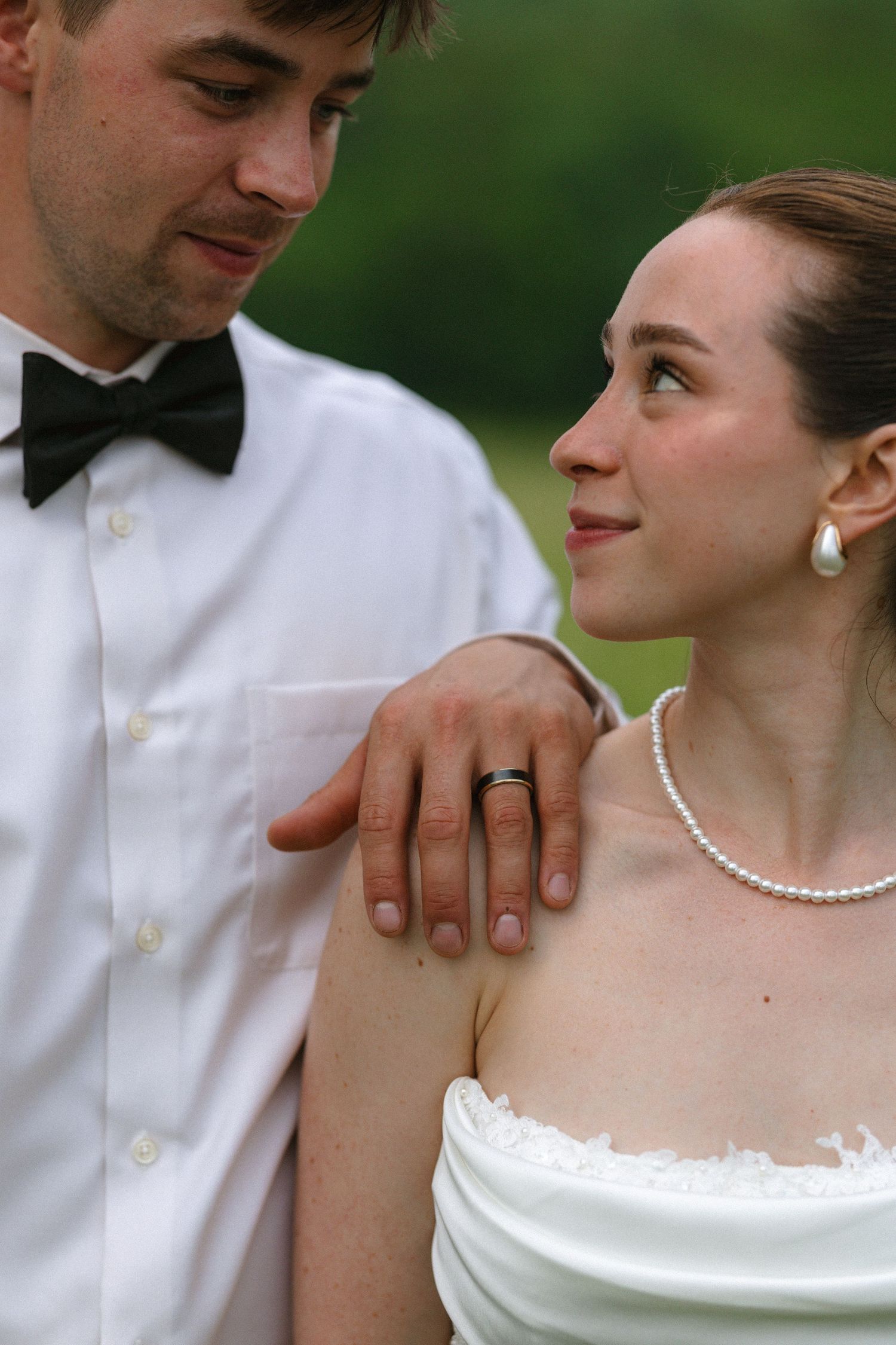 Groom with wedding ring rests arm on bride wearing pearl jewelry as they gaze at each other outdoors.