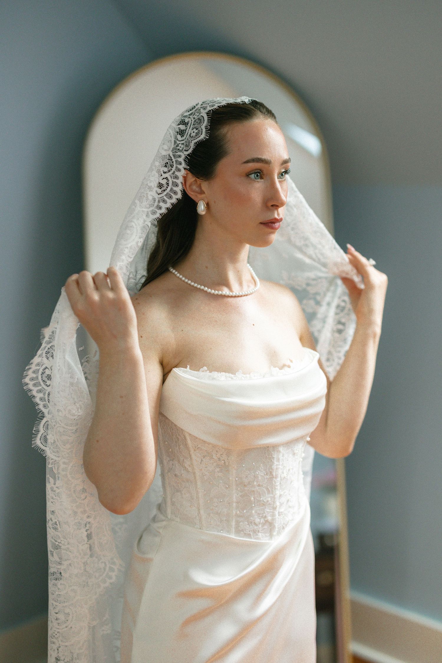 Bride in strapless lace wedding dress holding veil with pearl necklace standing before mirror.