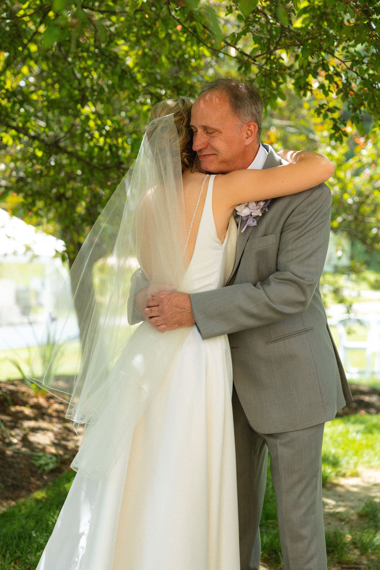 Bride in white gown and veil shares tender embrace with father in gray suit outdoors under green trees.