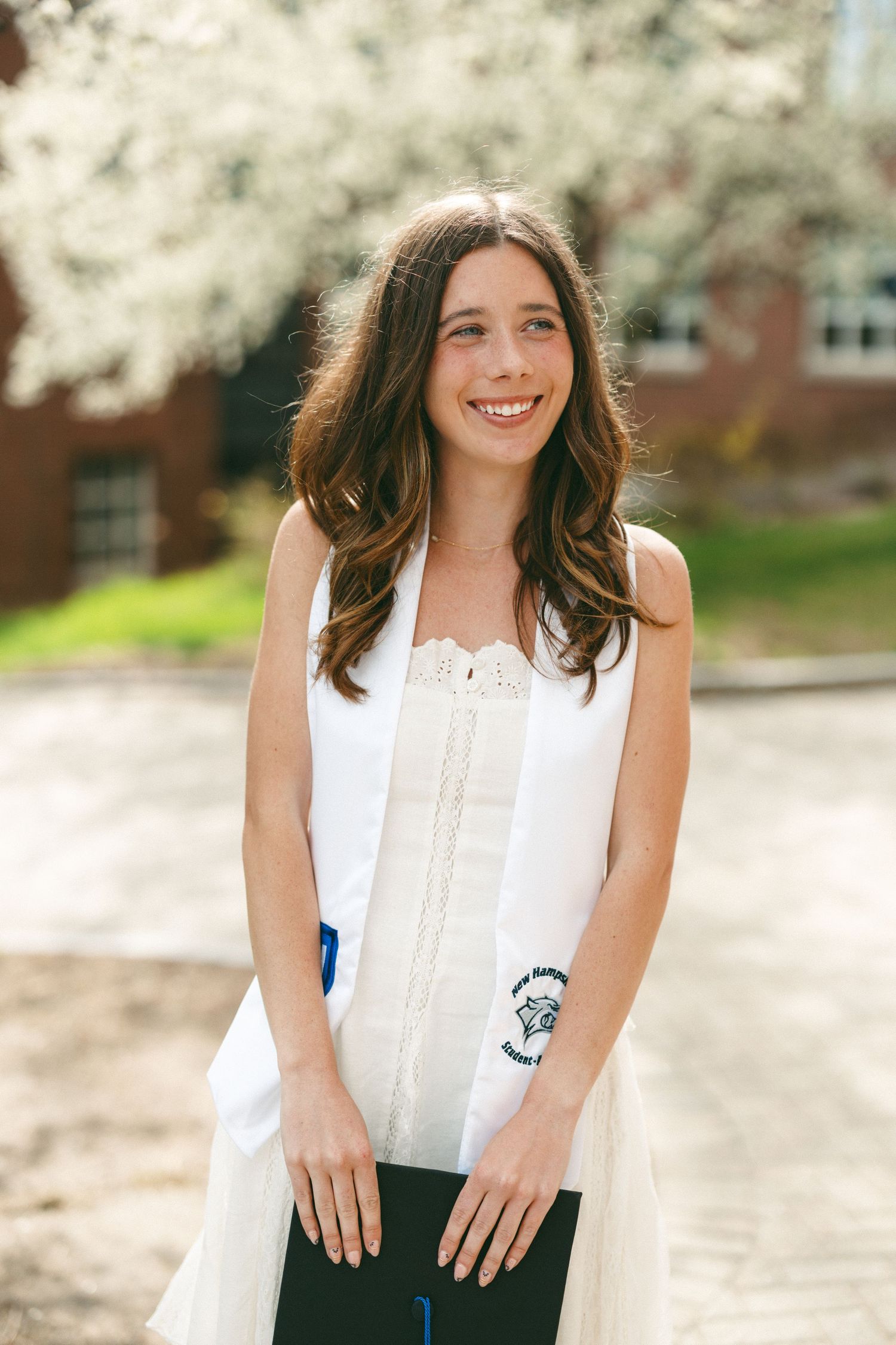 Smiling young woman in white graduation stole holding diploma outdoors near blooming trees.