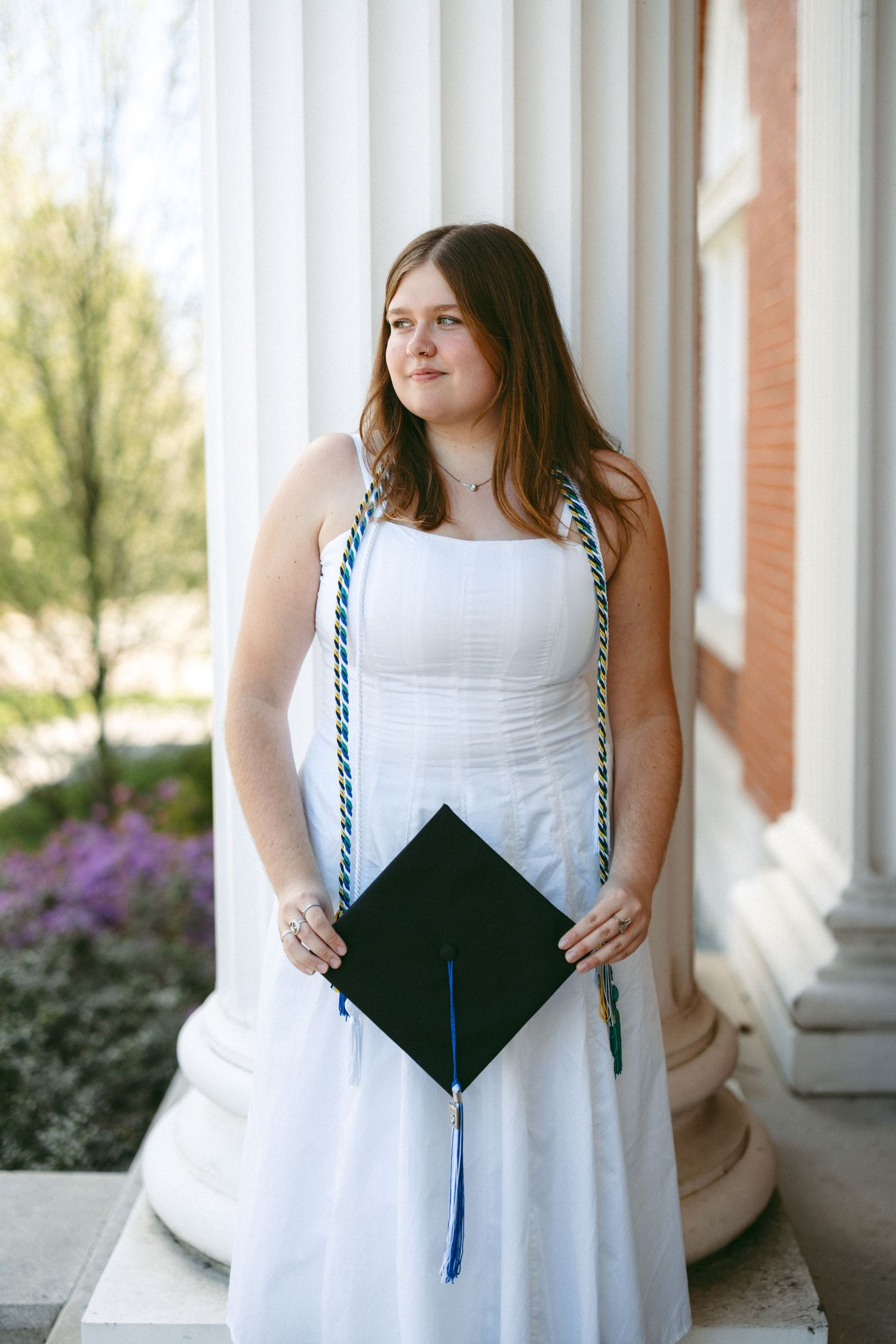Young woman in white dress holding graduation cap with honor cords, posing between white columns outdoors.