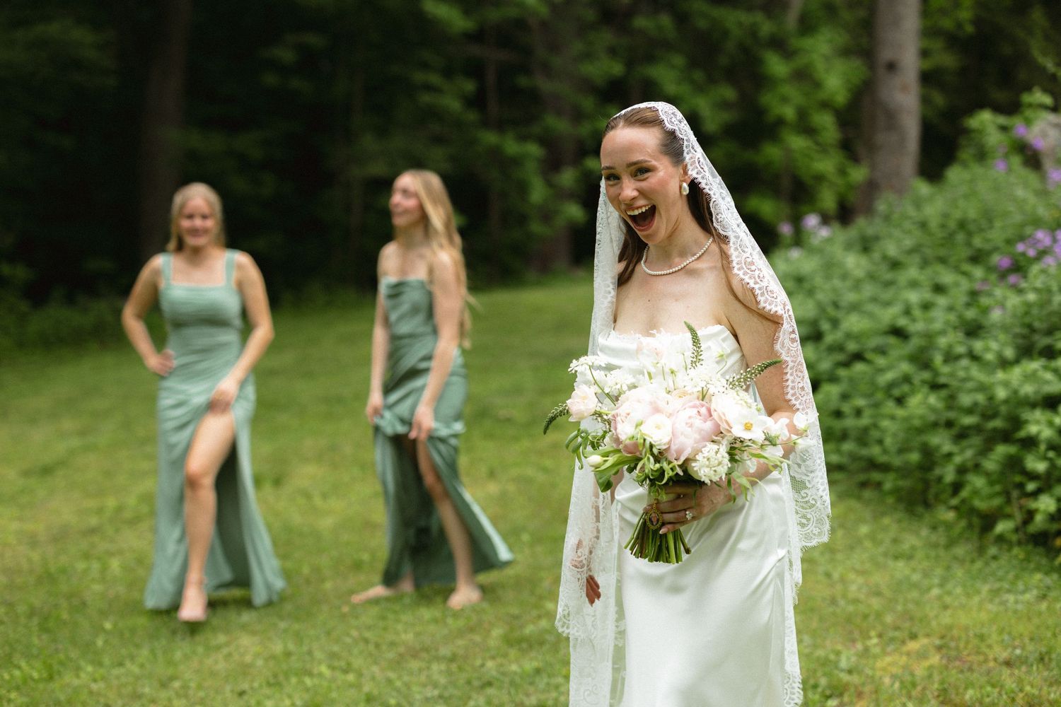 Laughing bride in white gown and veil holding bouquet with bridesmaids in sage green dresses behind her.