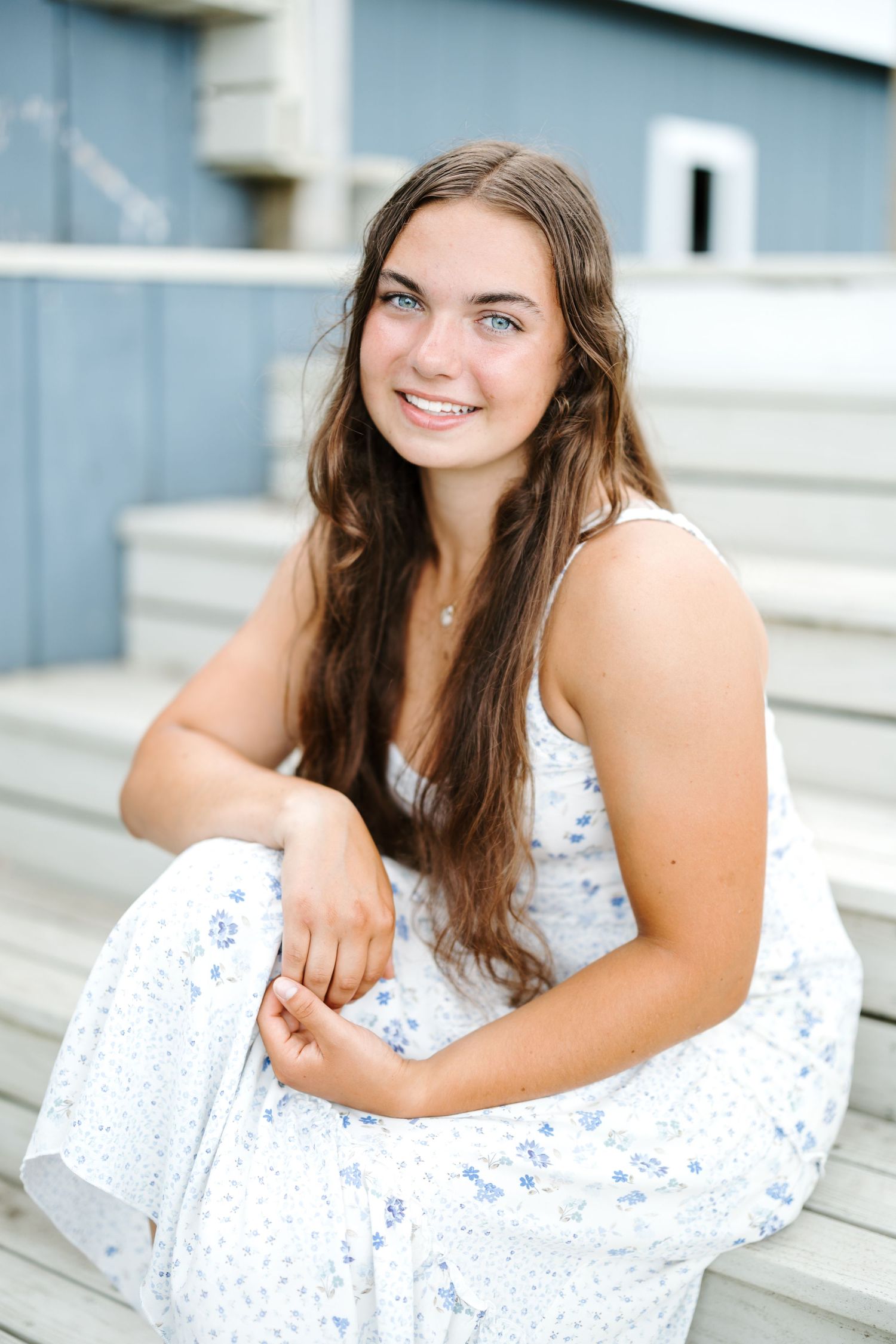 A senior in a white dress sits casually on outdoor stone steps during a portrait session.