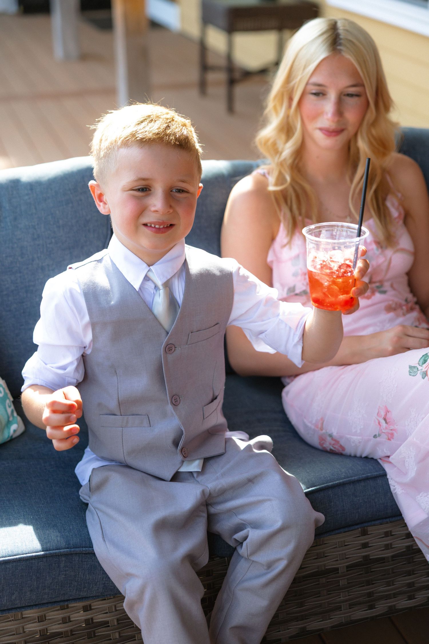 Young boy in gray suit smiles at camera while teen girl in pink dress holds iced drink at outdoor event.