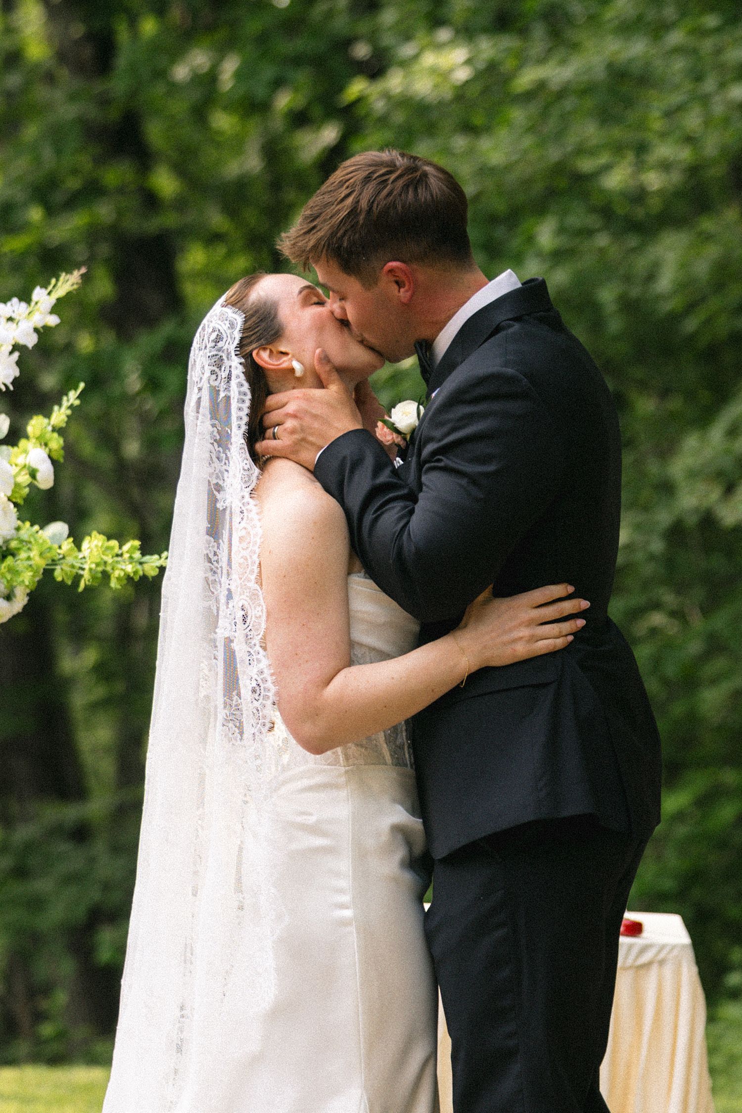 Bride and groom share their first kiss at an outdoor wedding ceremony surrounded by lush green trees.
