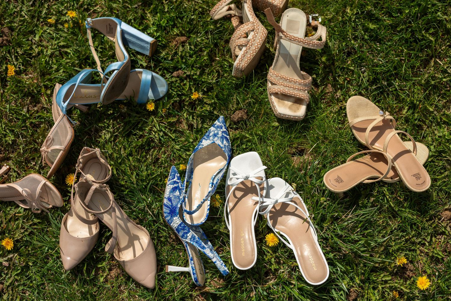 Assorted women's heels and sandals in nude, blue floral, and gold tones scattered on green grass.