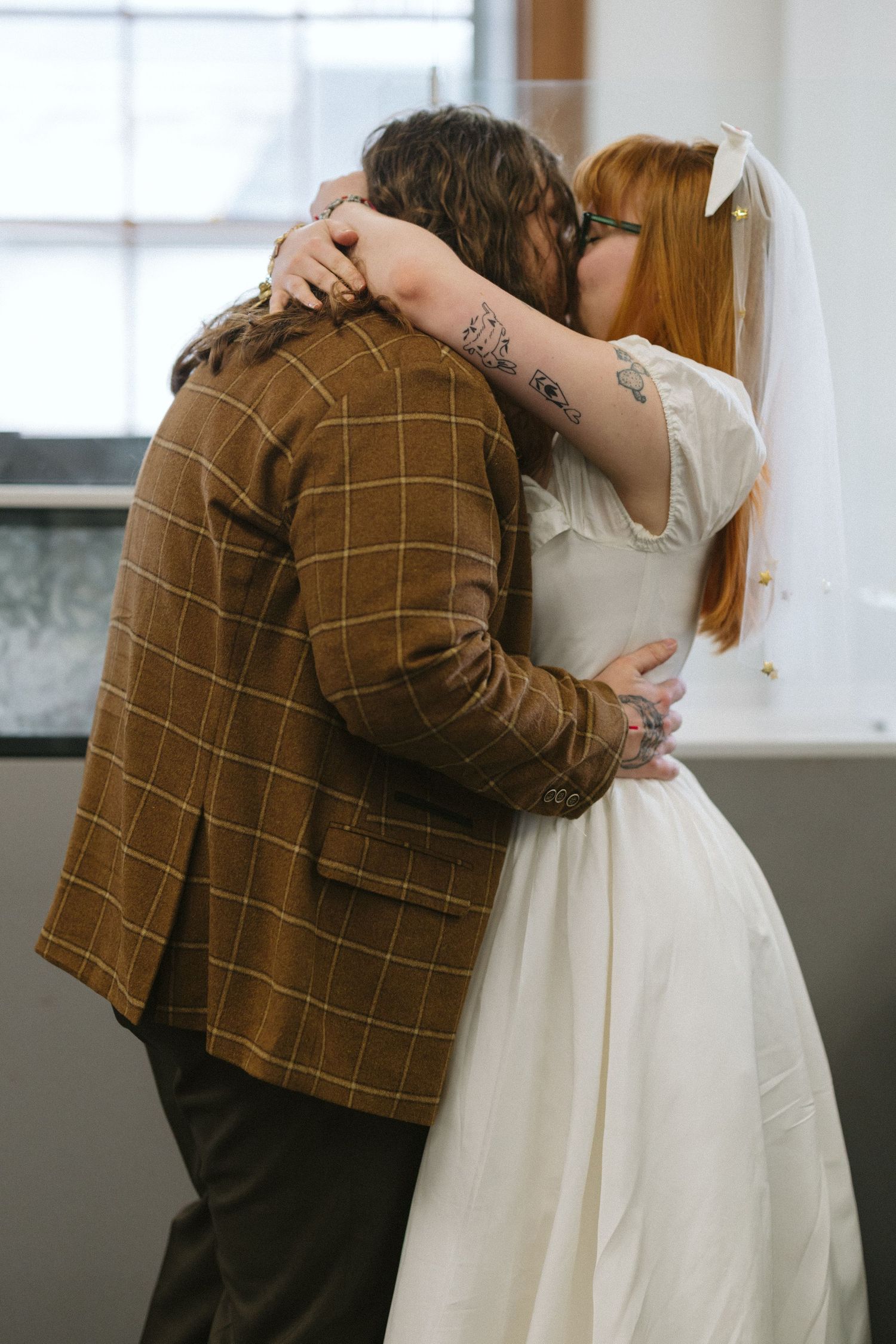 A bride in a white dress and veil embraces a groom in a brown plaid suit during their wedding ceremony.