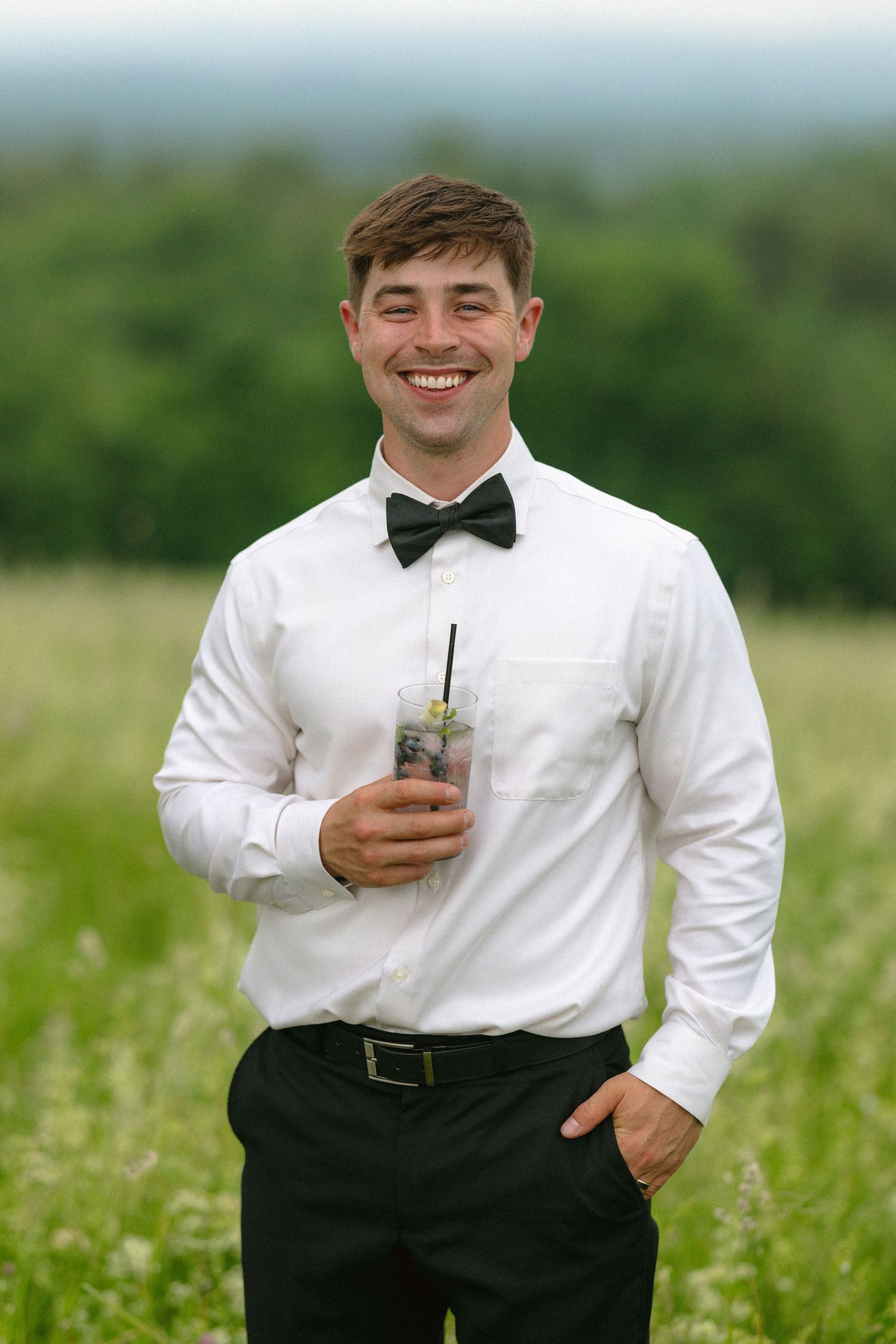 Smiling young man in white shirt and bow tie holding a cocktail drink in a green field outdoors.