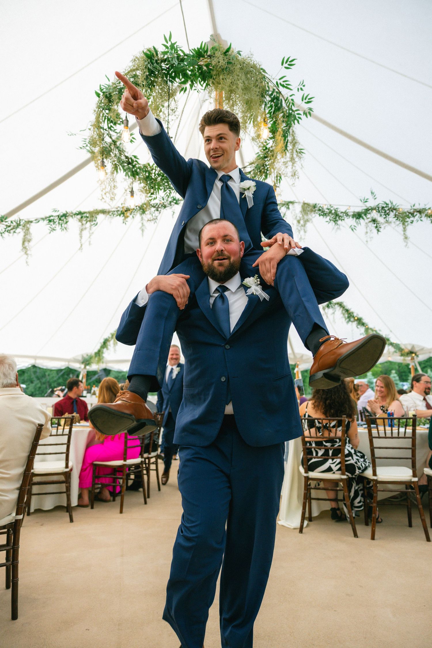 A groomsman rides on a guest's shoulders pointing ahead at a festive wedding reception under a tent.