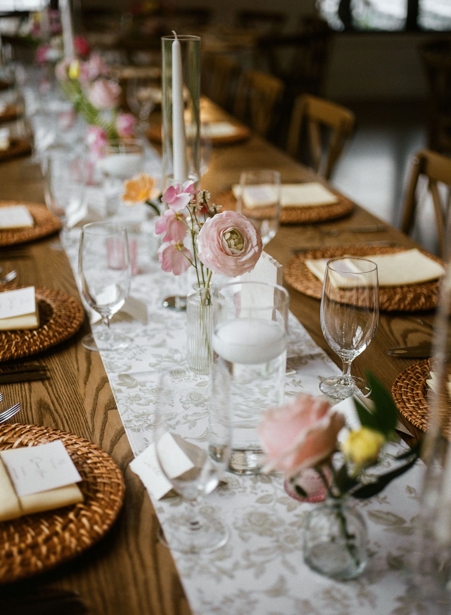 Elegant wedding reception table with pink roses, taper candles, woven chargers, and a floral lace runner on rustic wood.
