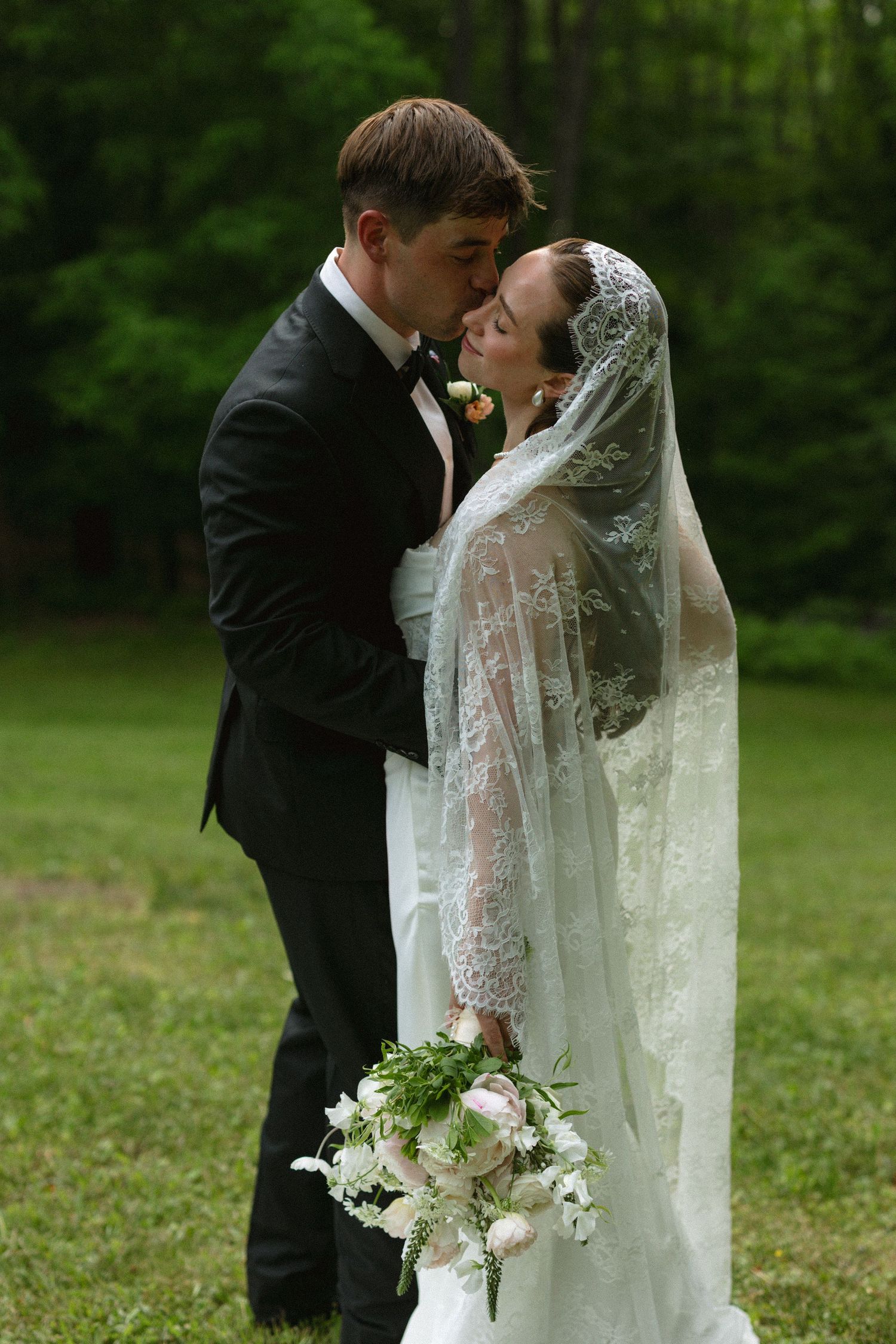 A bride and groom share a romantic kiss outdoors, with the bride wearing a lace veil and holding a bouquet.