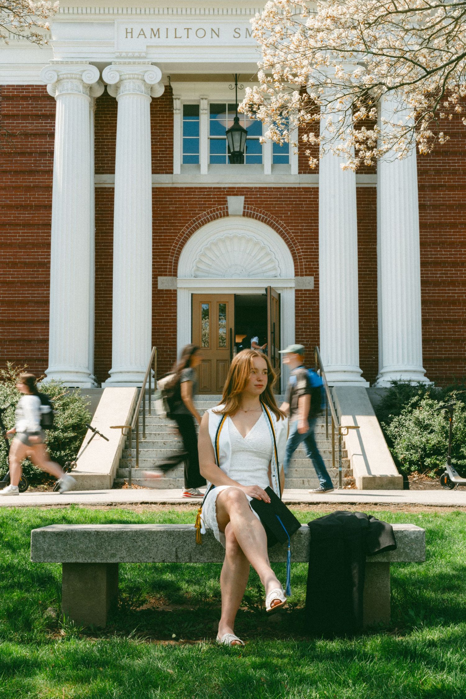 A student sits on a stone bench outside Hamilton Smith Hall, with blurred students passing by on a sunny spring day.