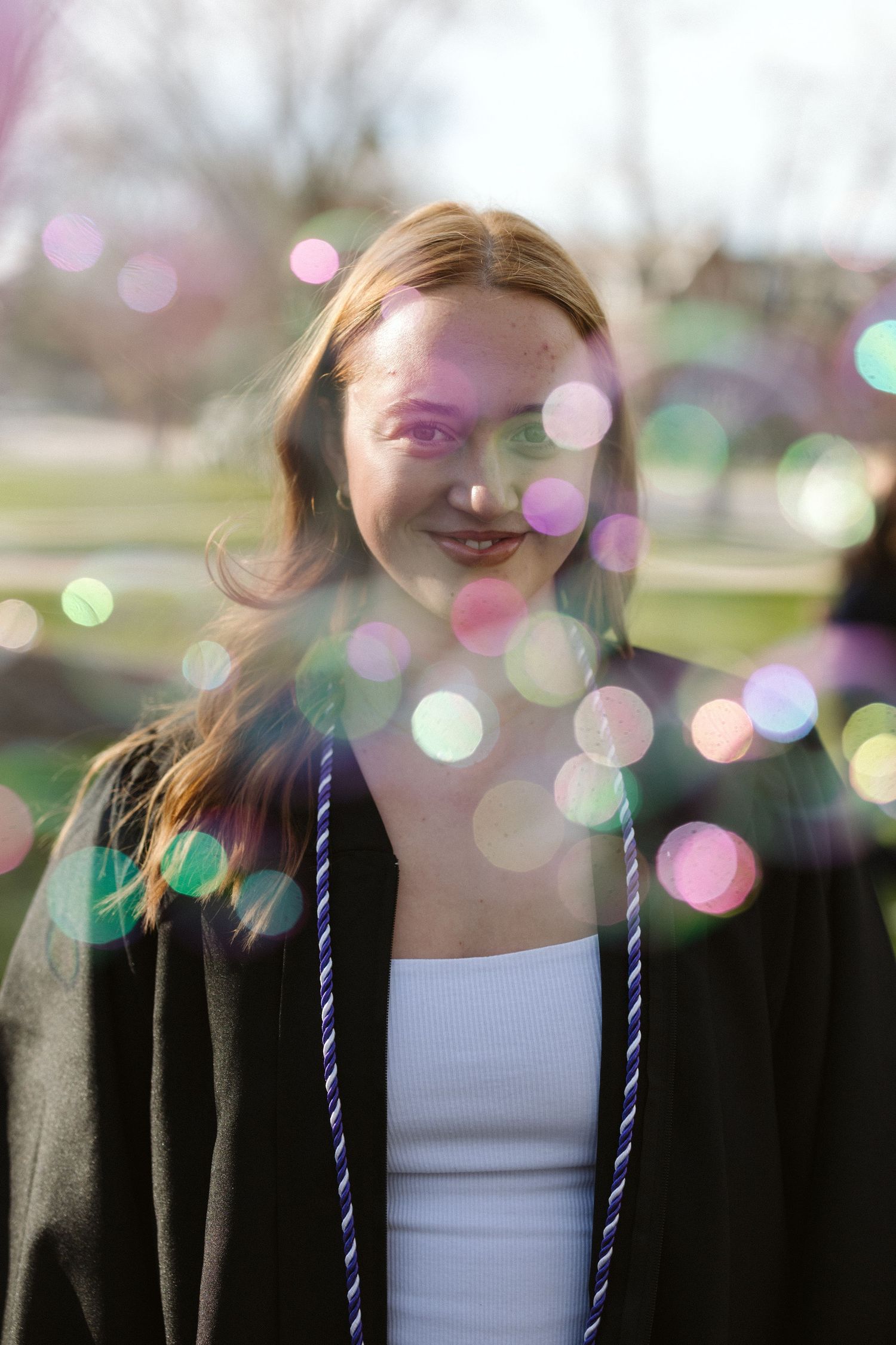 Young woman in graduation gown and honor cord smiles outdoors, surrounded by colorful bokeh light effects.