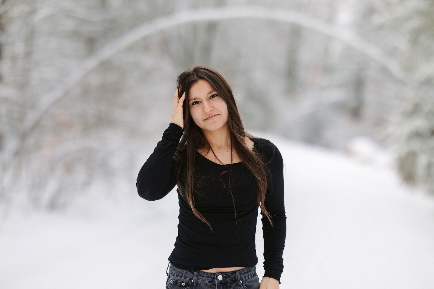 A person wearing a black sweater and jeans poses outdoors in a snowy winter landscape.