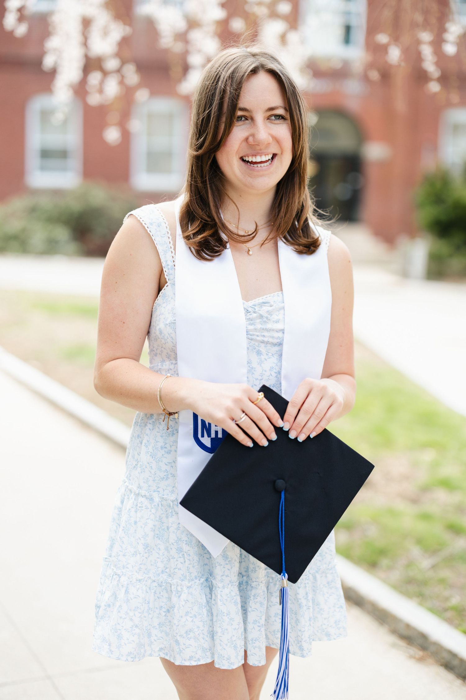 Graduate in white lace dress poses with graduation cap outdoors.
