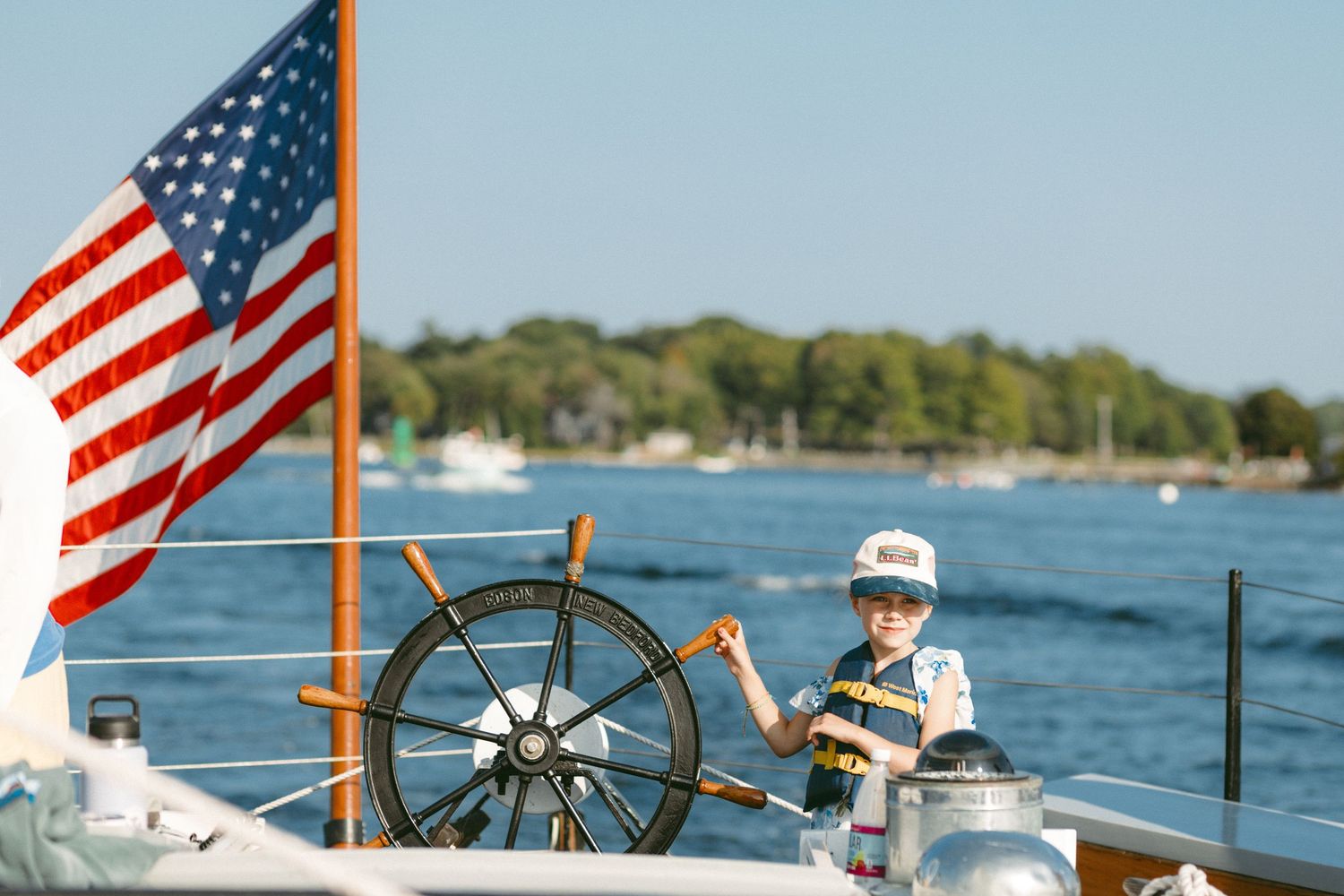 American flag waves near ship's wheel on a boat deck during sunny day.