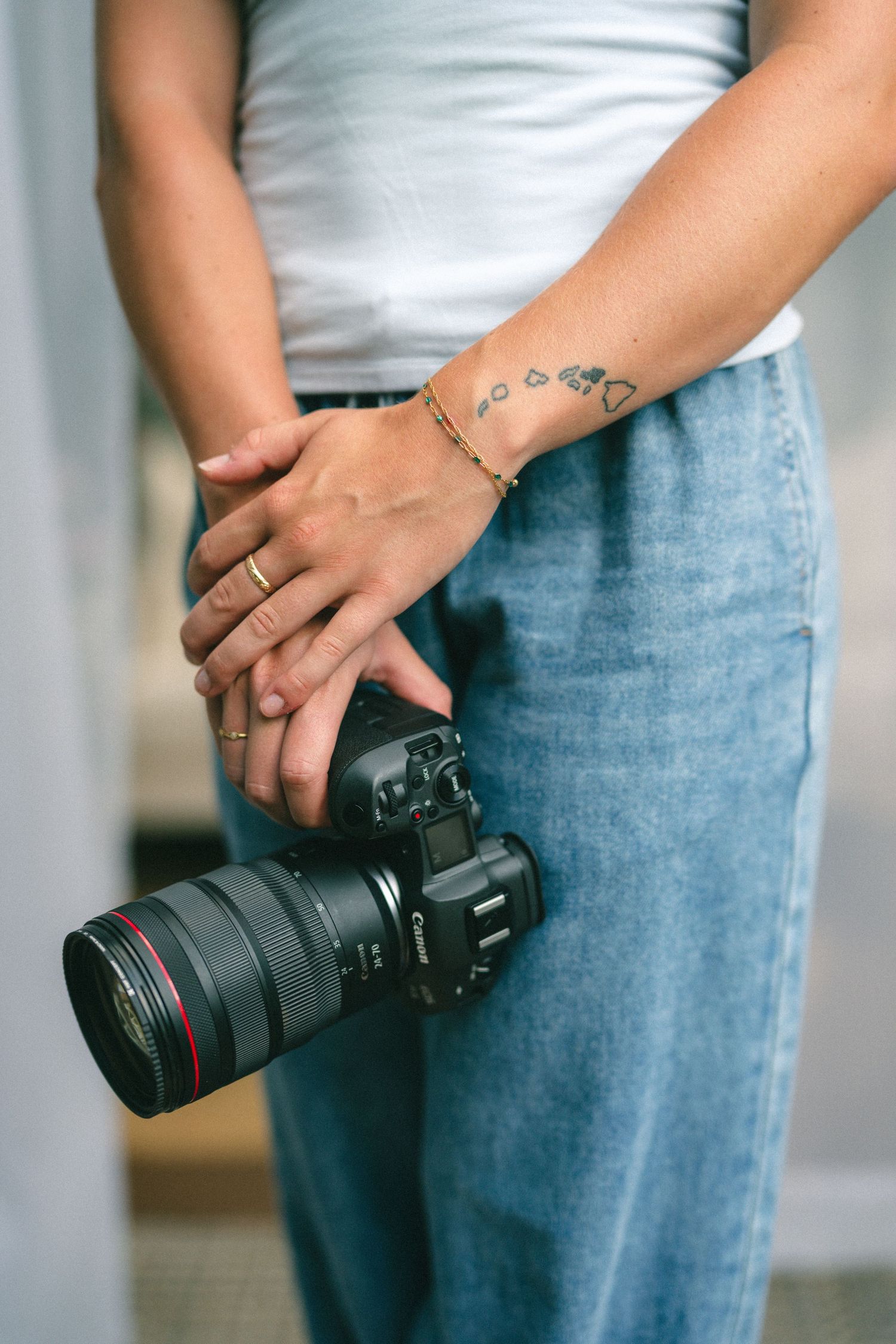 Person holding a Canon DSLR camera with tattoos and gold jewelry, wearing jeans and white top.