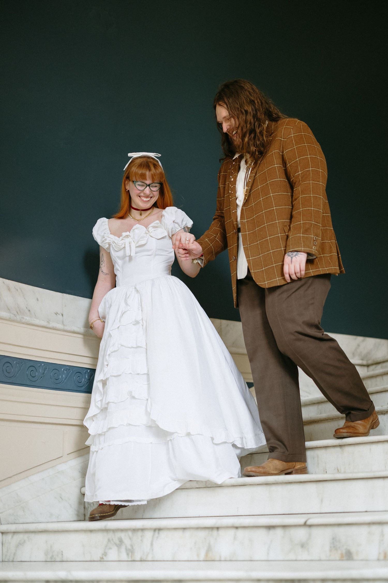 Redhead bride in white ruffled gown holds hands with long-haired groom in plaid jacket on marble stairs.