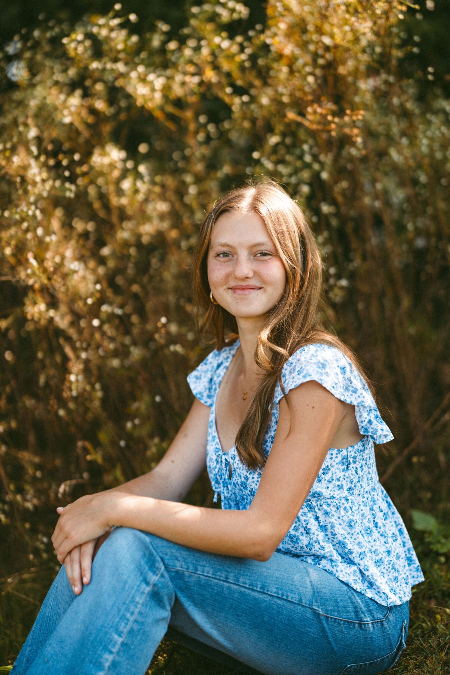 Young person in blue floral top poses outdoors against autumn foliage background.