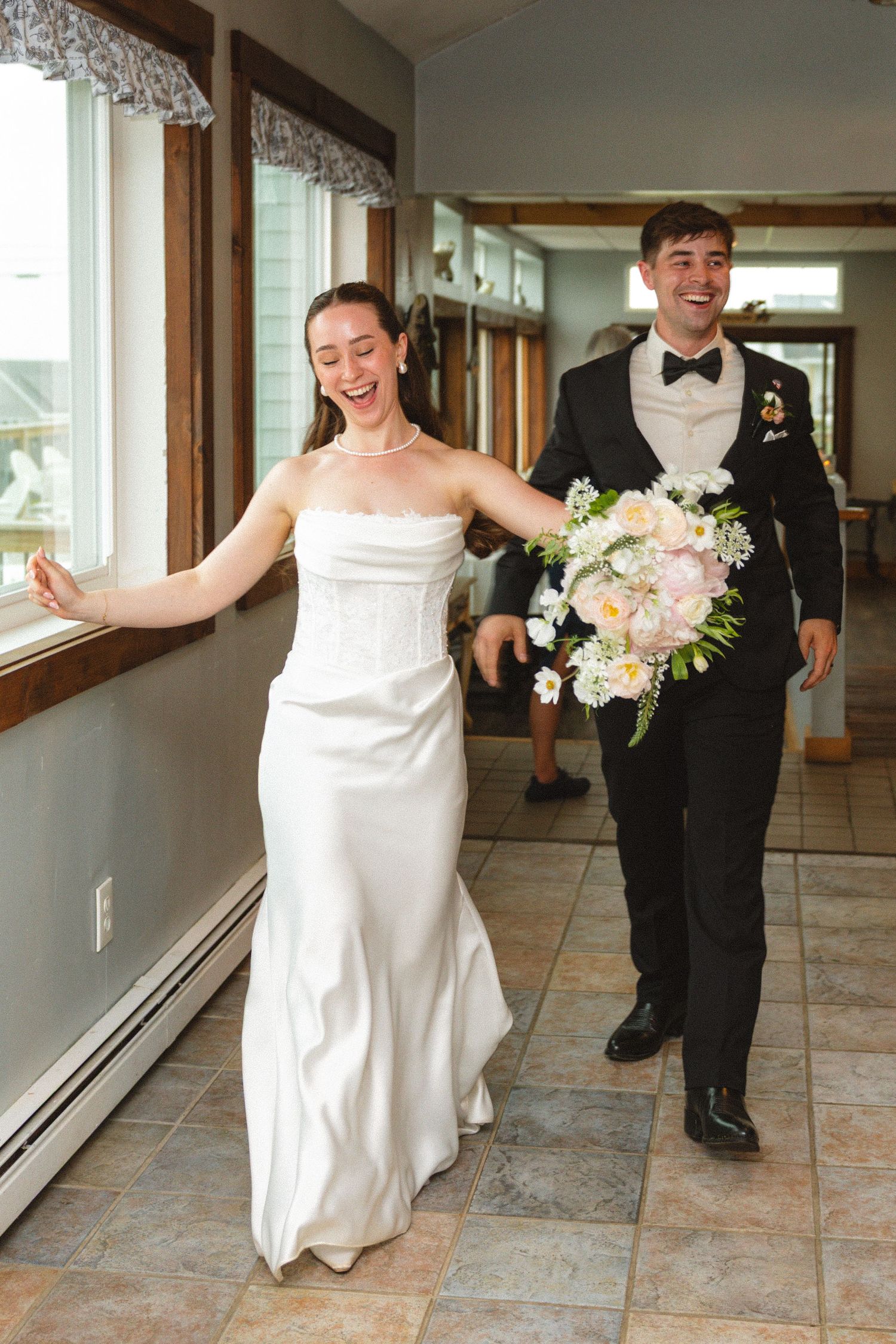 Joyful bride in strapless white gown and groom in tuxedo walking together holding a pastel bouquet indoors.