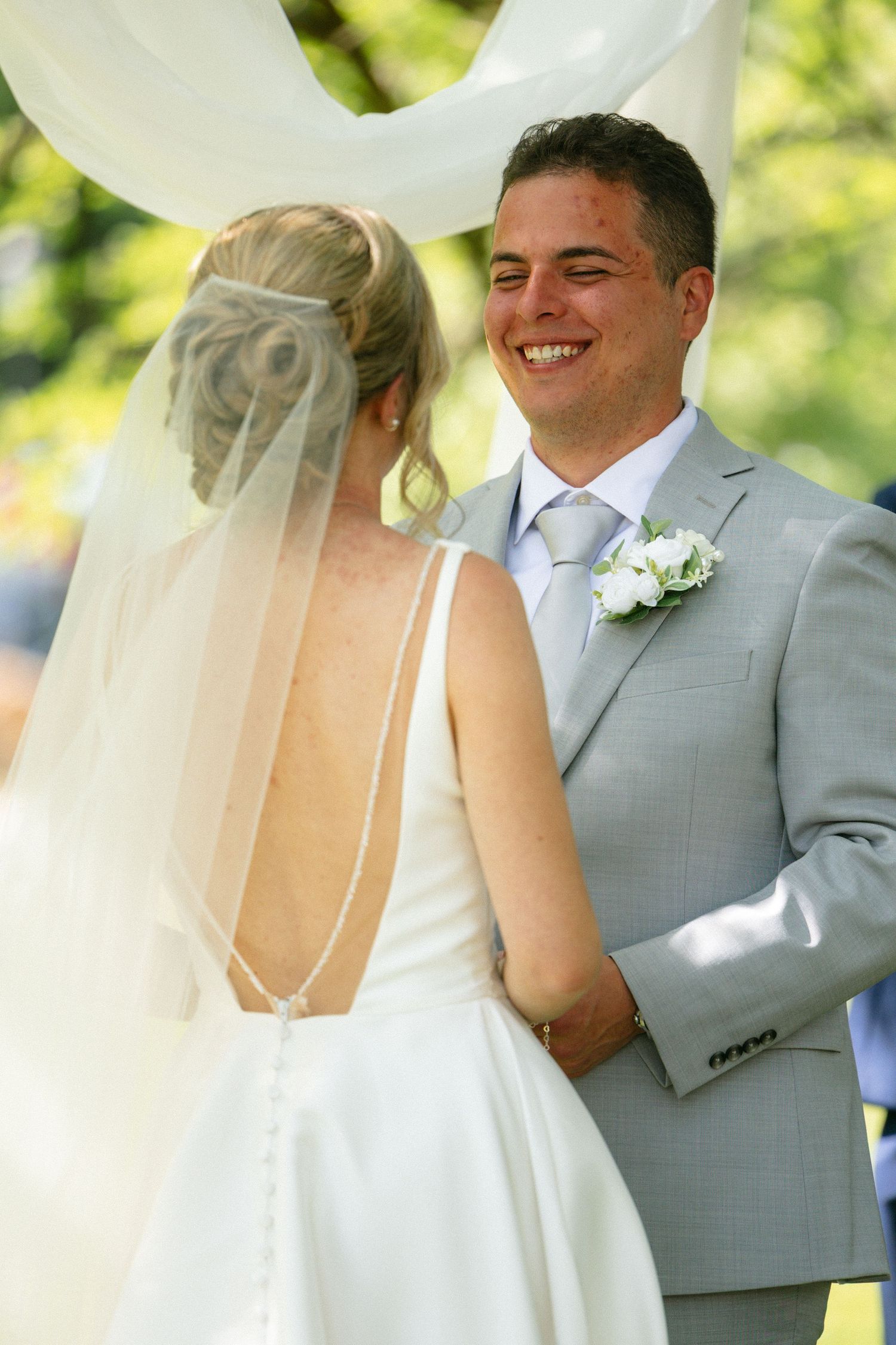 Smiling groom in gray suit with white boutonniere faces bride in backless wedding dress with veil at outdoor ceremony.