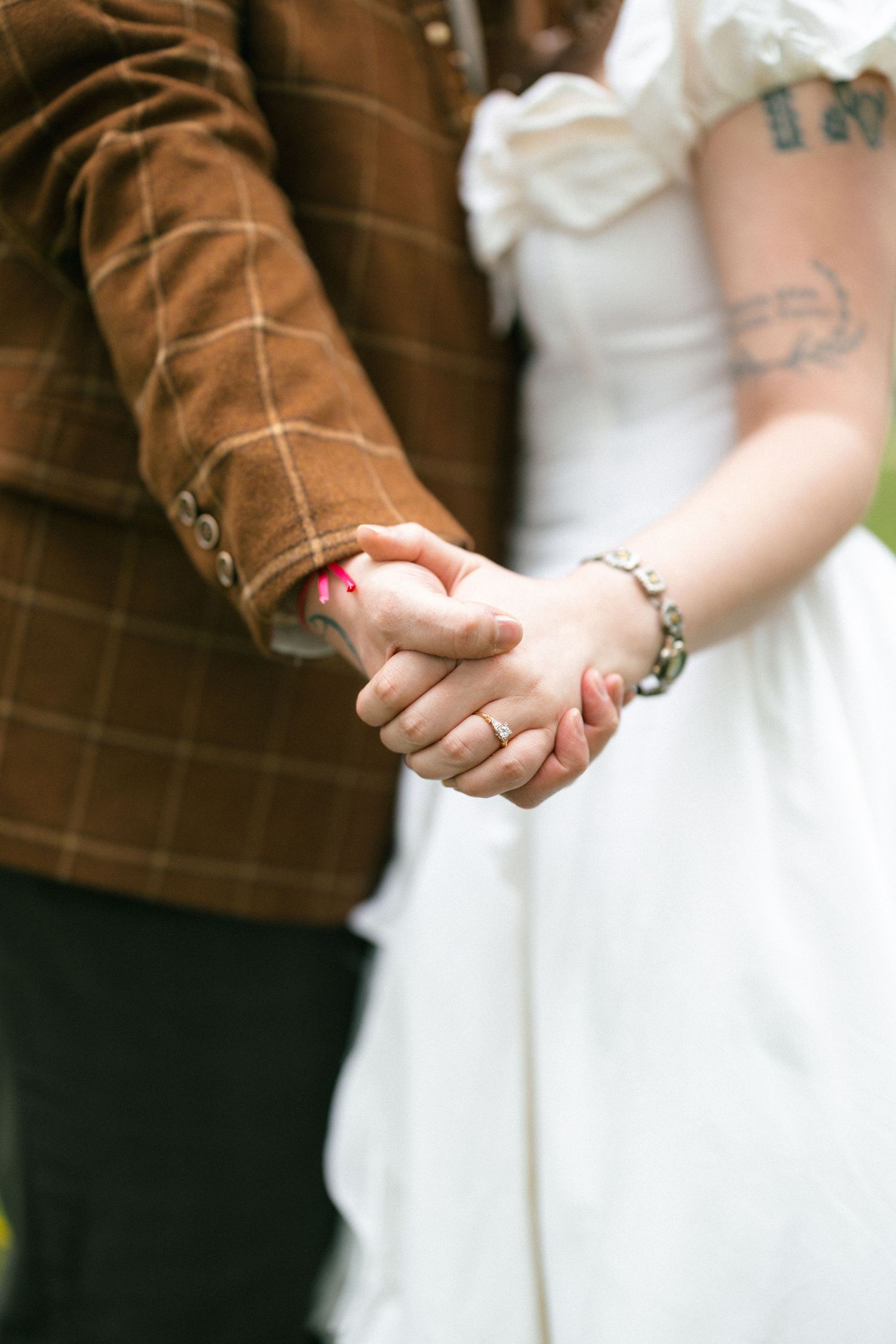 A bride and groom hold hands, showcasing wedding rings, a beaded bracelet, and a tattooed arm in elegant attire.