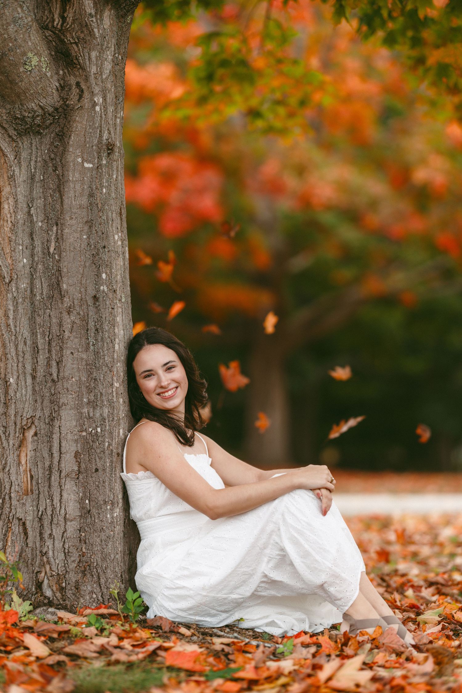 A white dress flows gracefully among fallen autumn leaves beneath tall trees with vibrant orange foliage.