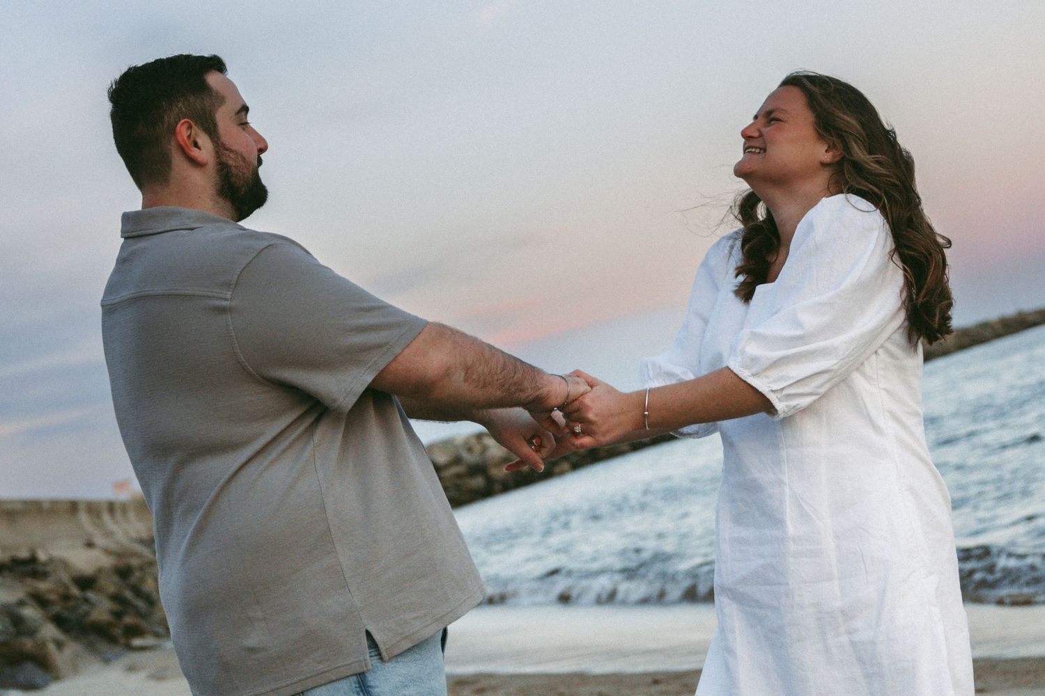 Couple holding hands on rocky beach at sunset, woman in white dress and man in gray shirt smiling romantically.
