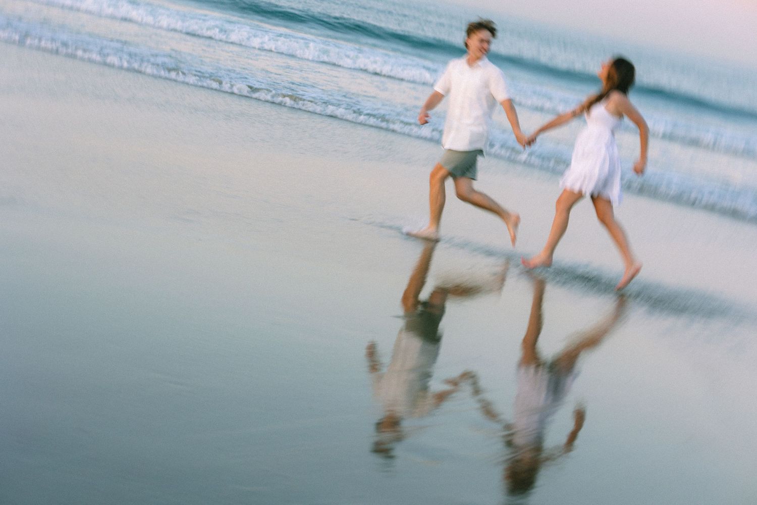 Couple holding hands running on a wet beach at sunset, wearing white outfits with reflections on the sand.