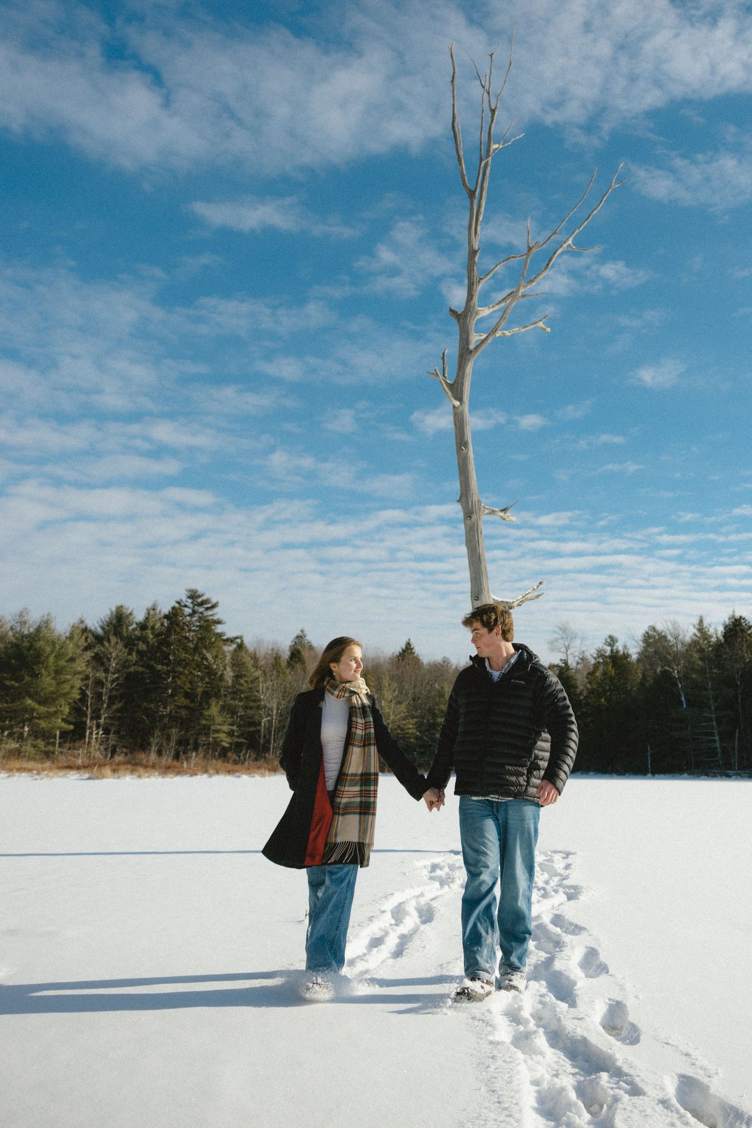 A couple walks hand in hand across a snowy field on a bright winter day with bare trees and blue skies overhead.