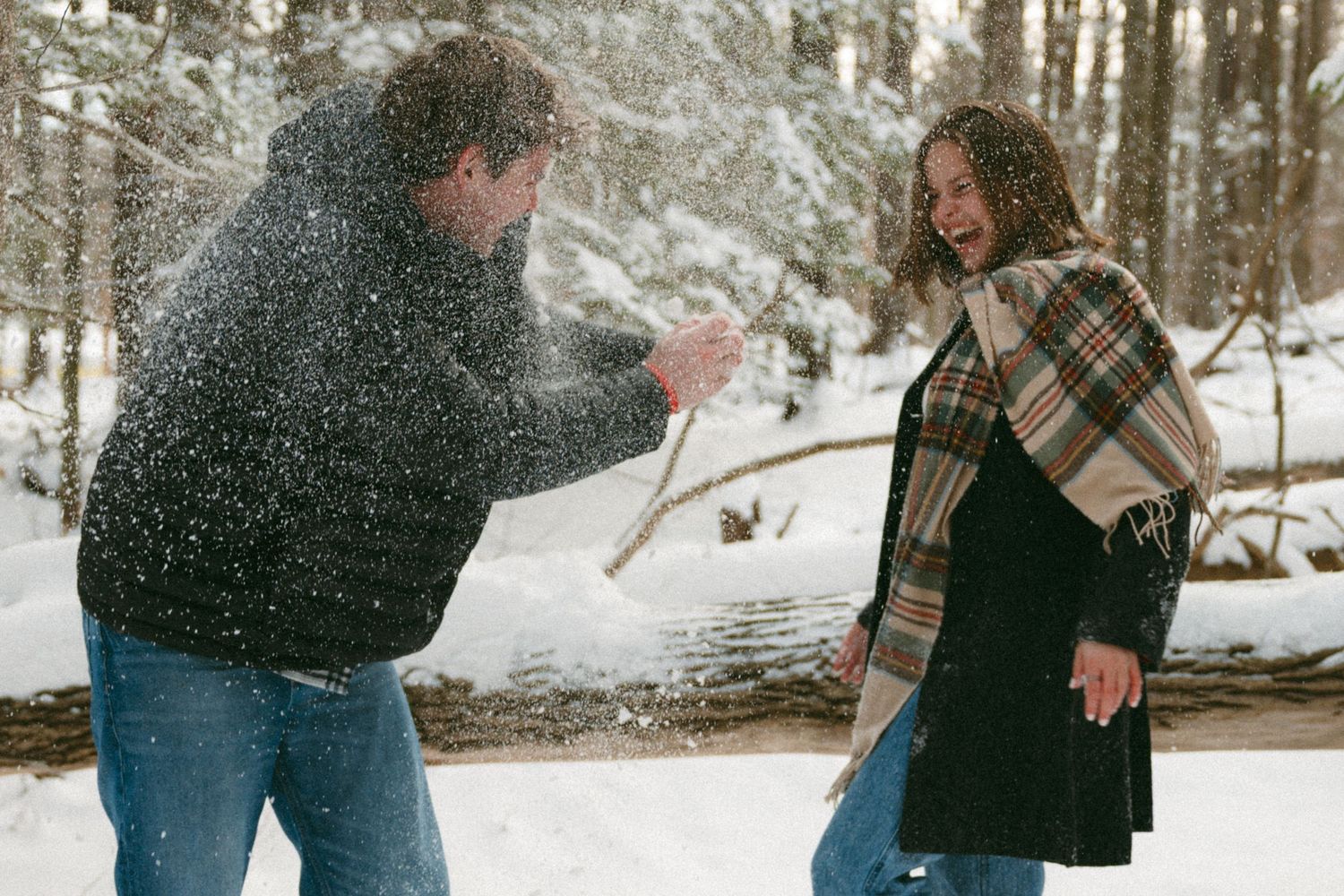 A couple stands together in a snowy winter forest setting during a romantic outdoor photoshoot.