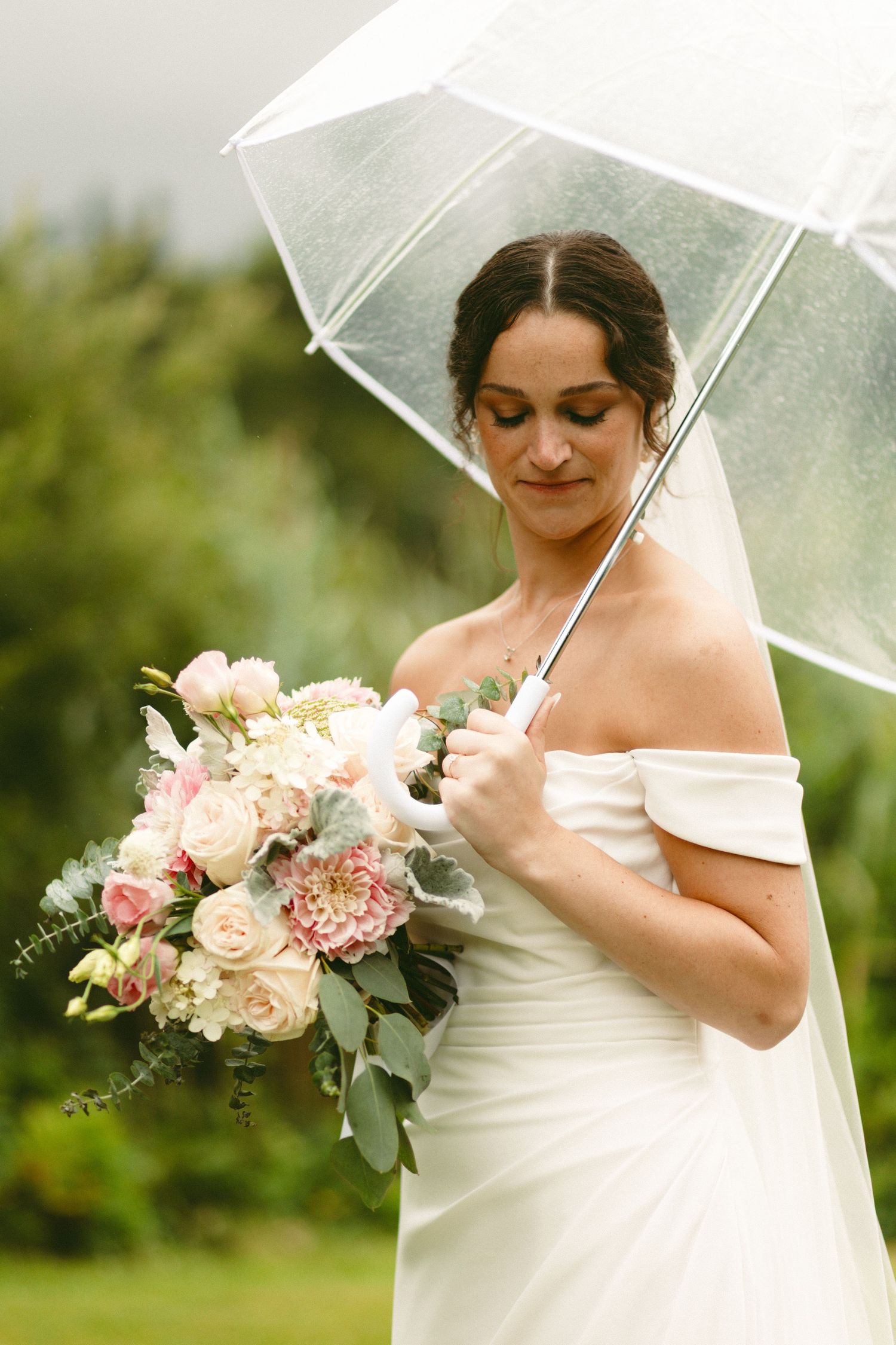 Bride in white dress holding clear umbrella with pink and white floral bouquet.