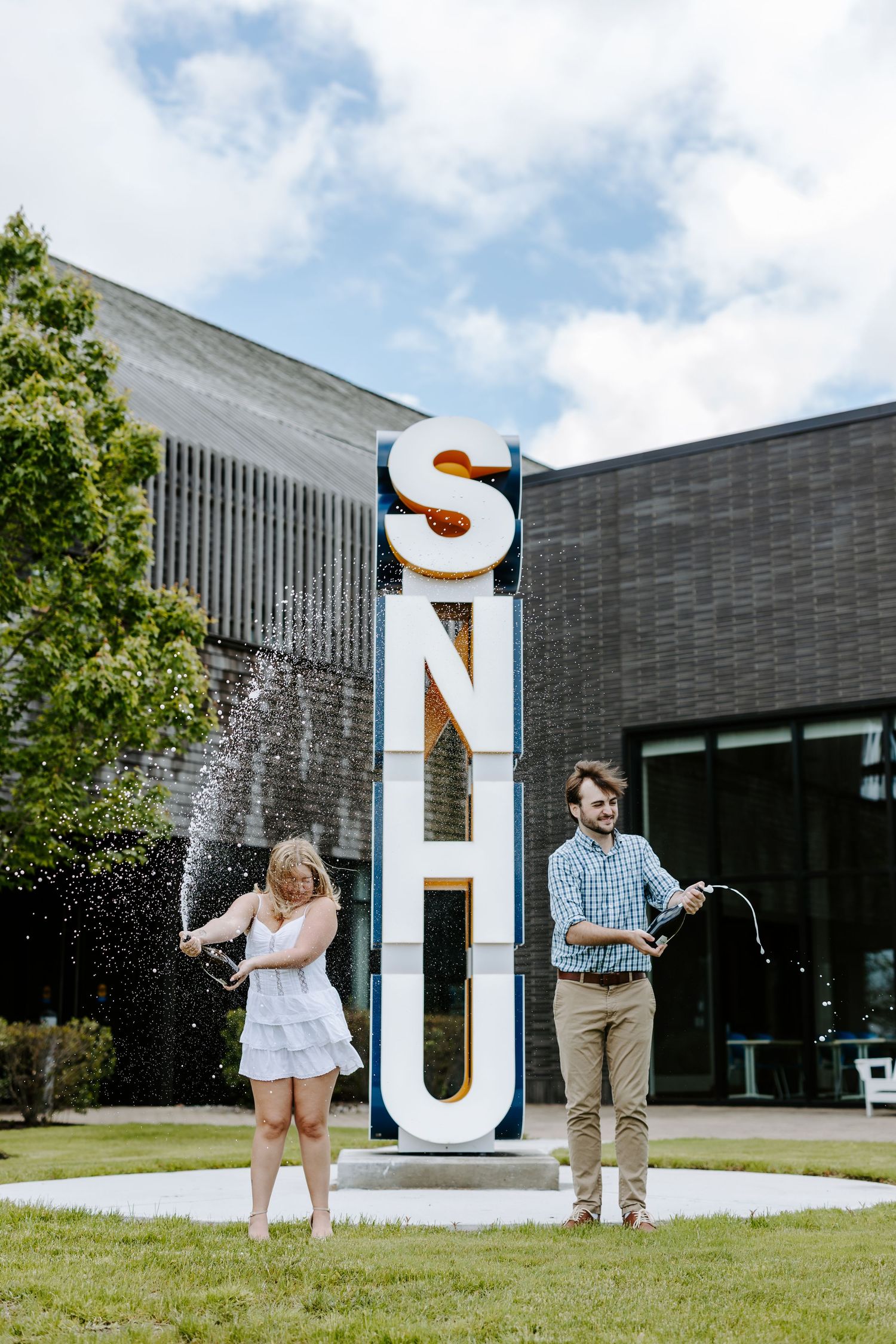 Graduates posing with large UNH letters on campus grounds with water splashing.