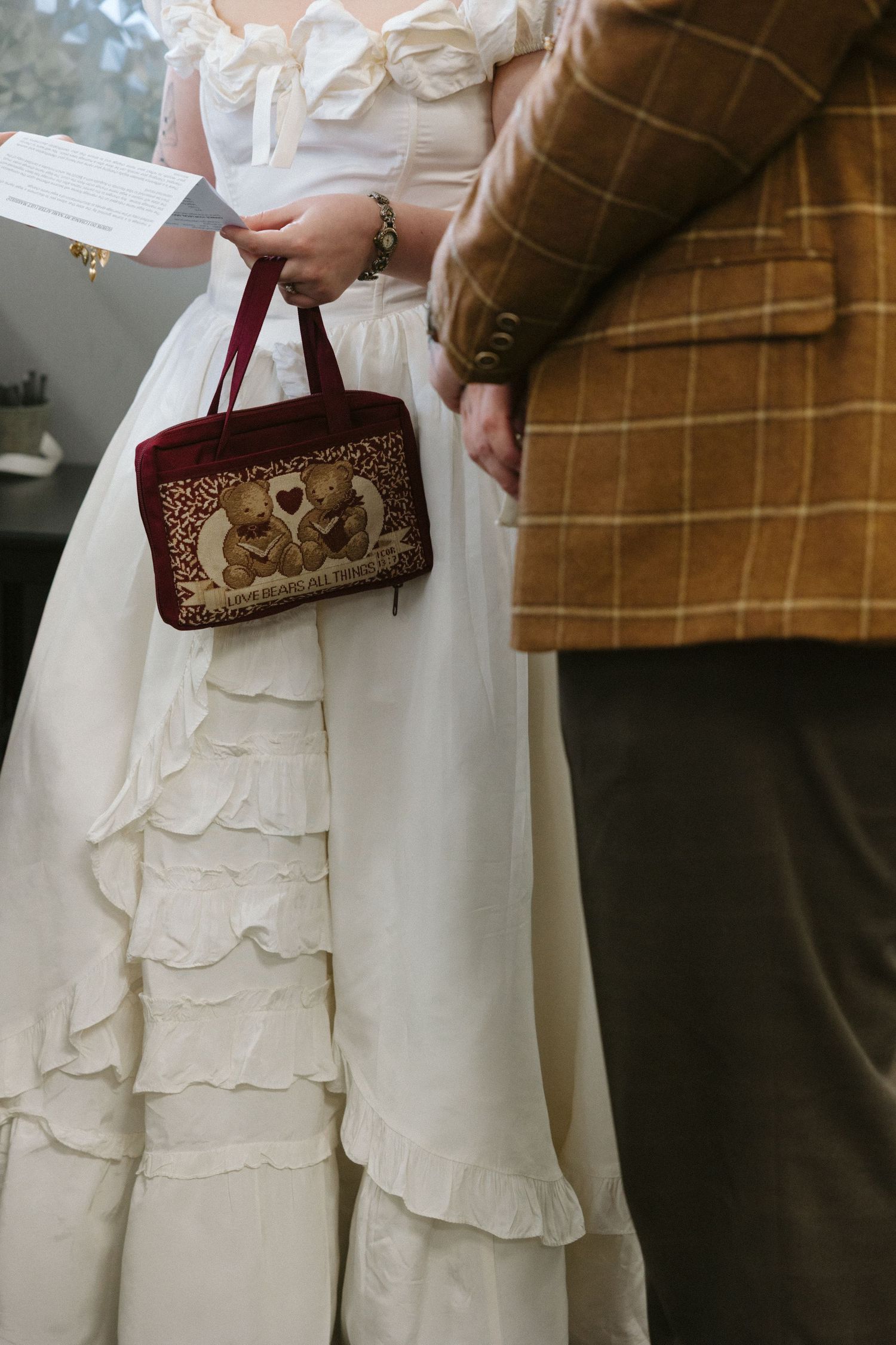 Bride in ruffled white gown holding velvet bear-embroidered handbag beside groom in plaid suit at wedding ceremony.