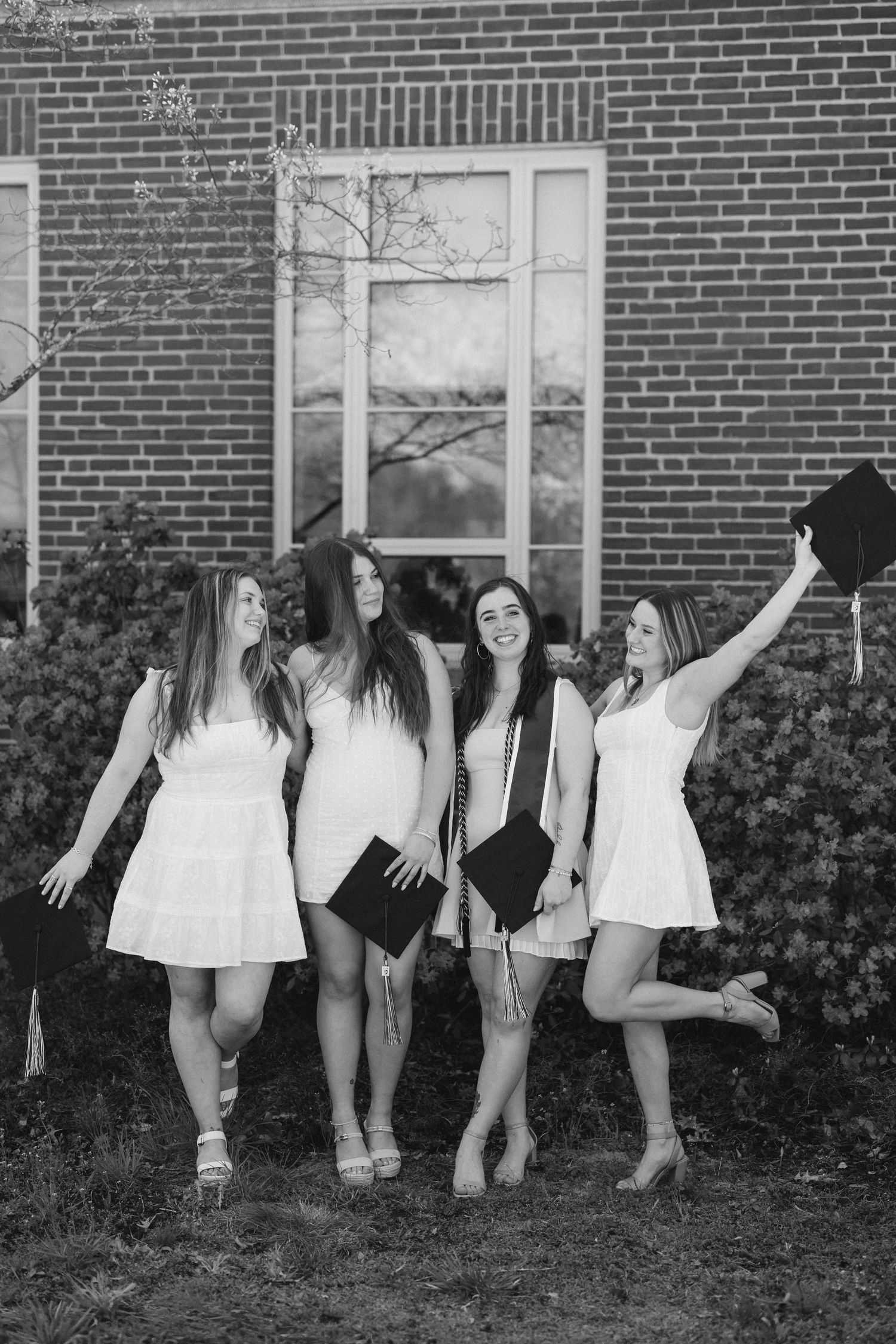 Four young women in white dresses celebrate graduation, holding caps outdoors near a brick building in black and white photo.