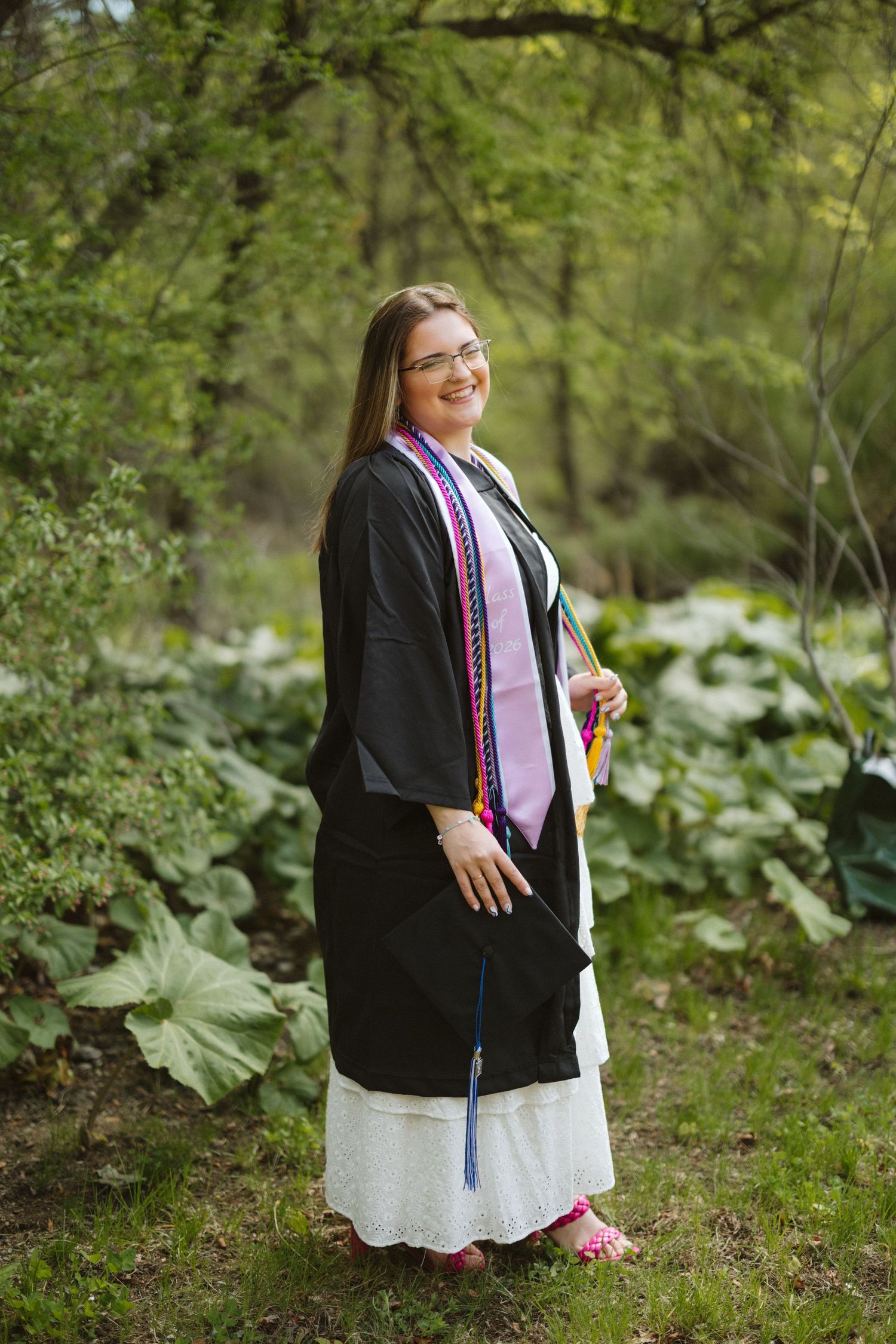 A smiling graduate in black gown and colorful stoles poses outdoors among lush green trees and plants.