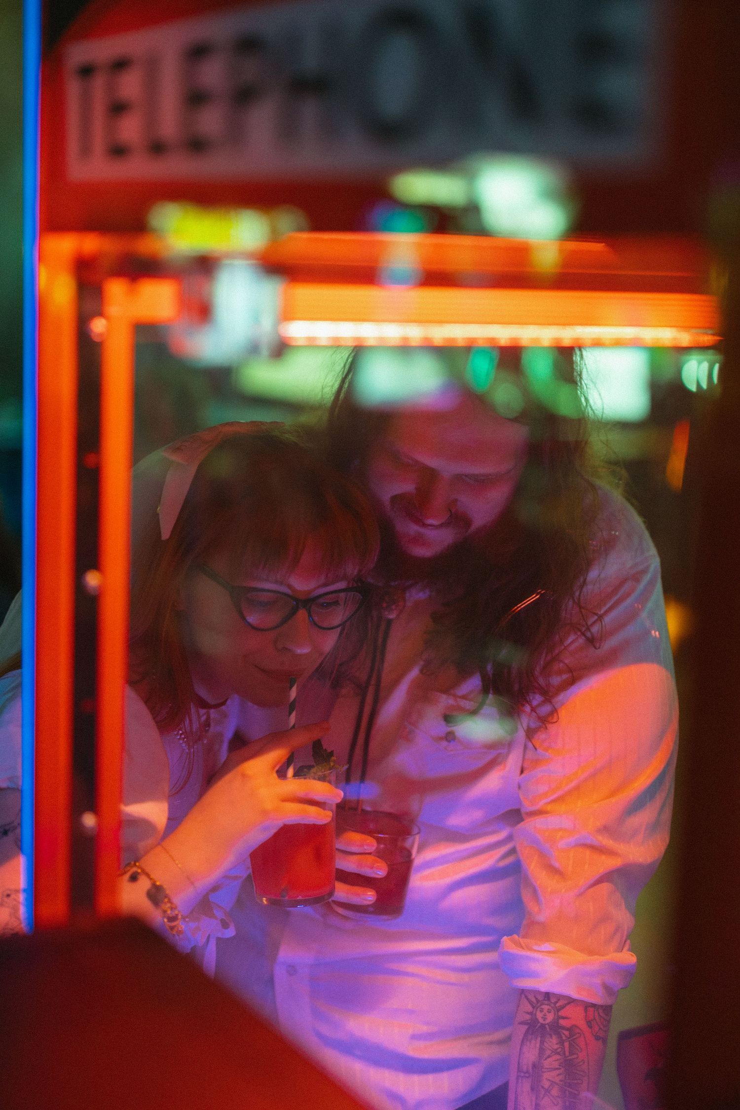 Couple sharing a moment inside a neon-lit telephone booth, smiling at a phone screen at night.