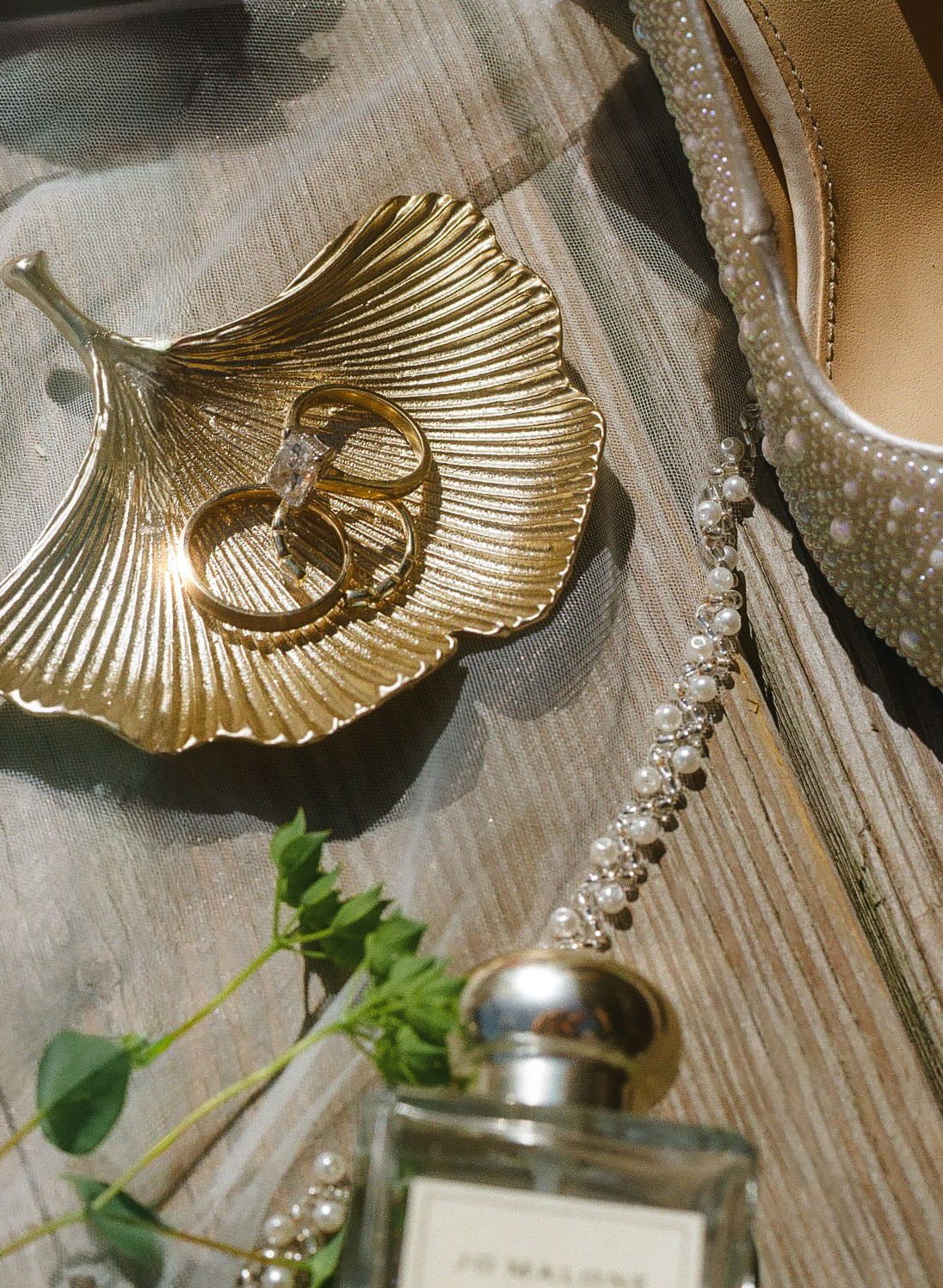 Gold leaf dish with wedding rings, pearl necklace, crystal heels, and Jo Malone perfume on wooden surface.