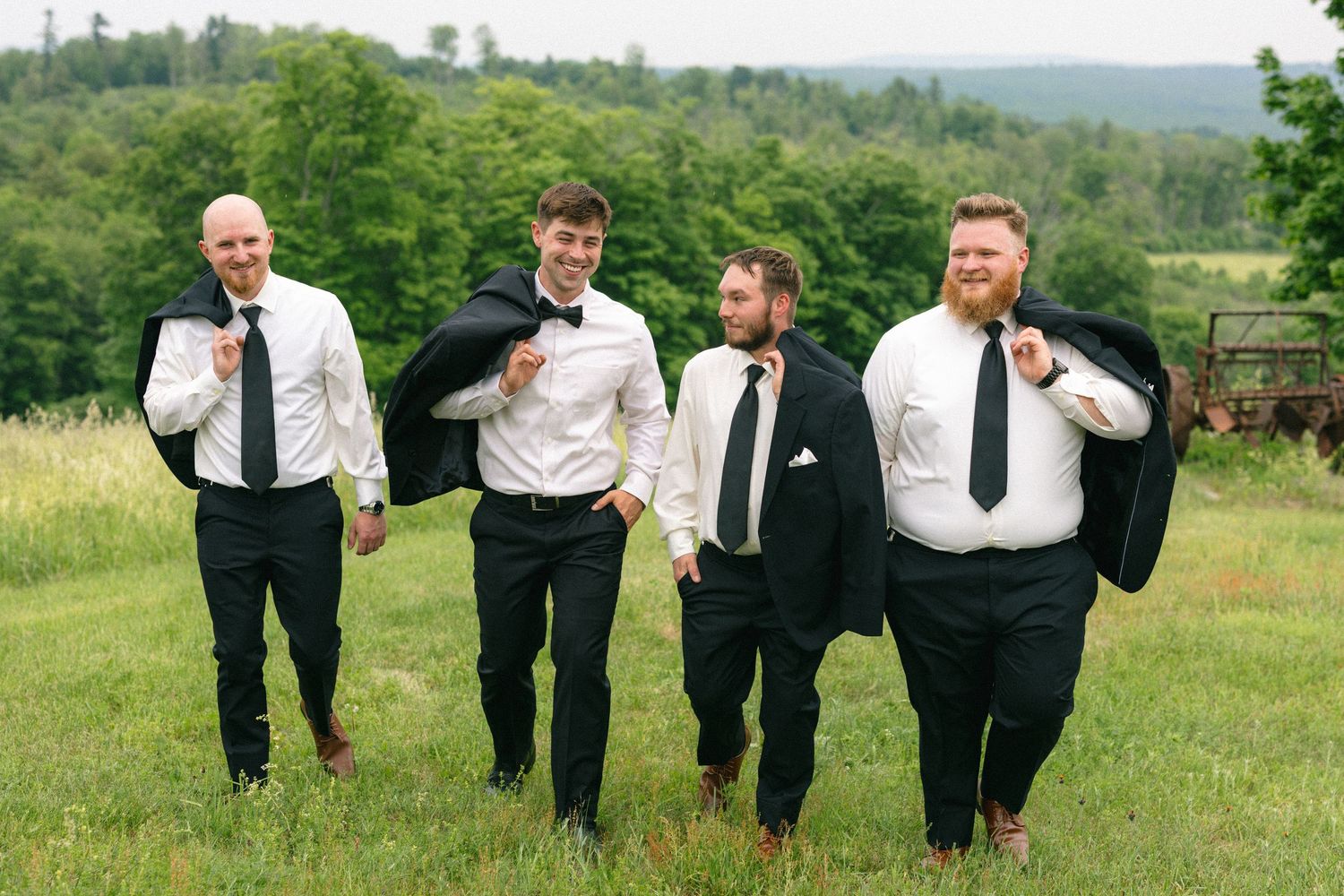 Four groomsmen in black suits and ties walking outdoors in a green field on a wedding day.