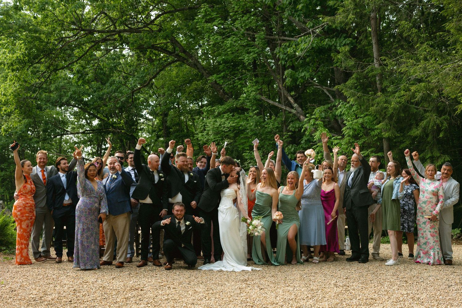 Large wedding party celebrating outdoors with raised hands among lush green trees on gravel pathway.