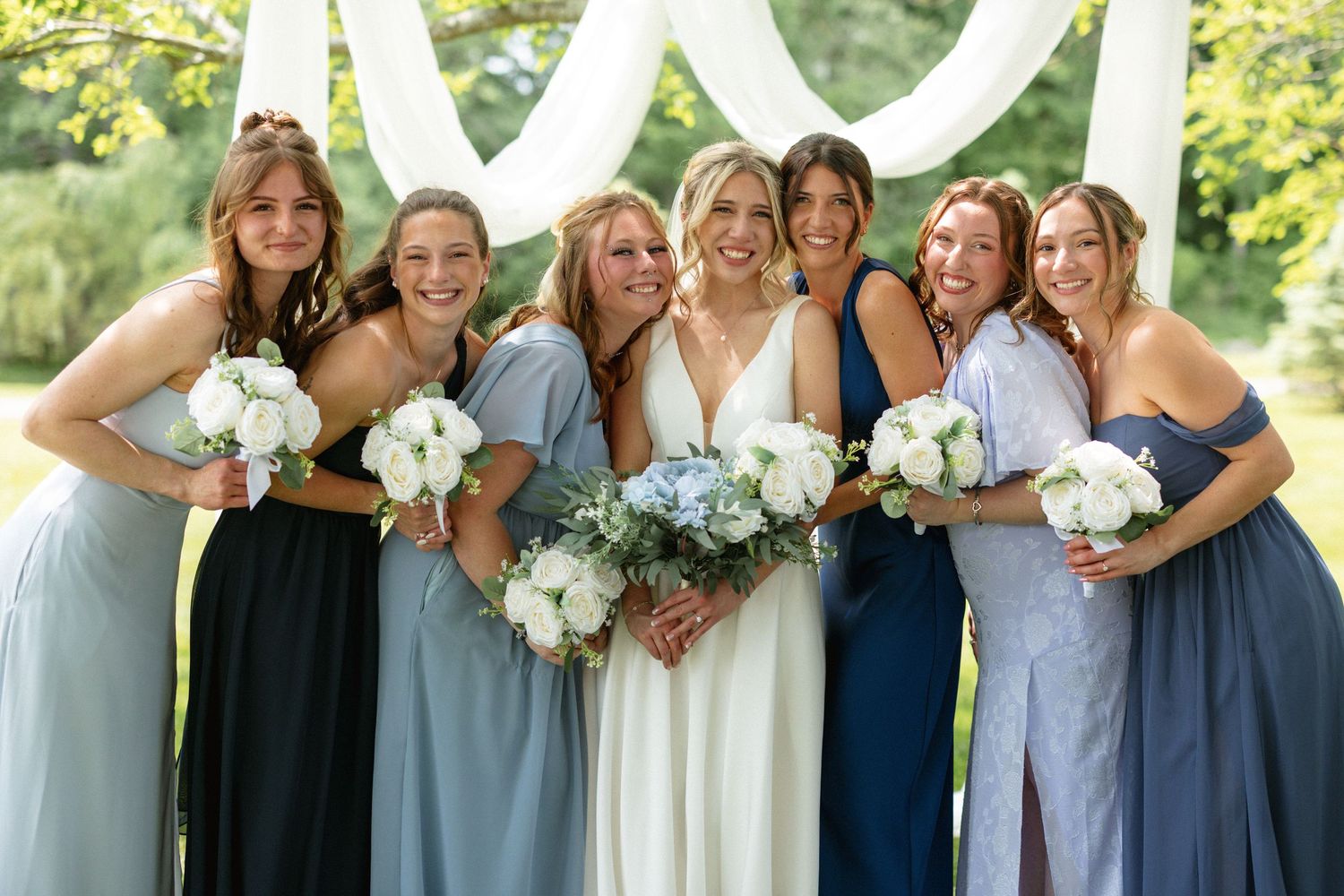 Smiling bride in white gown poses with six bridesmaids in blue dresses holding white bouquets outdoors.